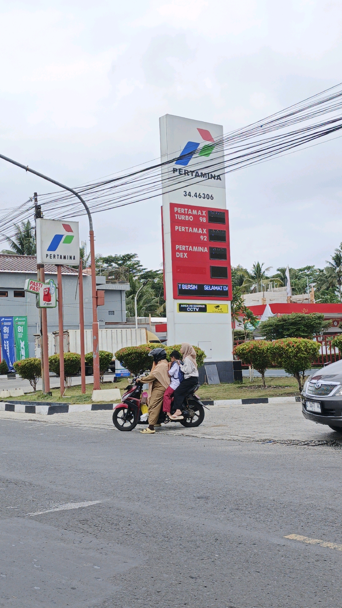A Pertamina gas station is seen in Desa Karangjaladri, Indonesia, under a cloudy sky. A tall sign displays Pertamina branding and fuel prices: Pertamax Turbo 98, Pertamax 92, and Pertamina Dex. Below the prices, a digital display reads "T BERSIH SELAMAT DA" and an illuminated yellow sign states "AREA INI DIAWASI CCTV". Two Pertamina signs are visible, one with the Pertamina logo and another smaller sign with a cartoon character saying "PASTI PAS!". In the foreground, a red motorcycle carries three passengers: an adult male driver, an adult female passenger behind him, and a child in front of the adult female. The passengers are dressed casually. The motorcycle is on the asphalt road. To the right, a grey car is parked, showing its front grille and license plate "L 1677 WQ". The area is lined with green bushes and trees.