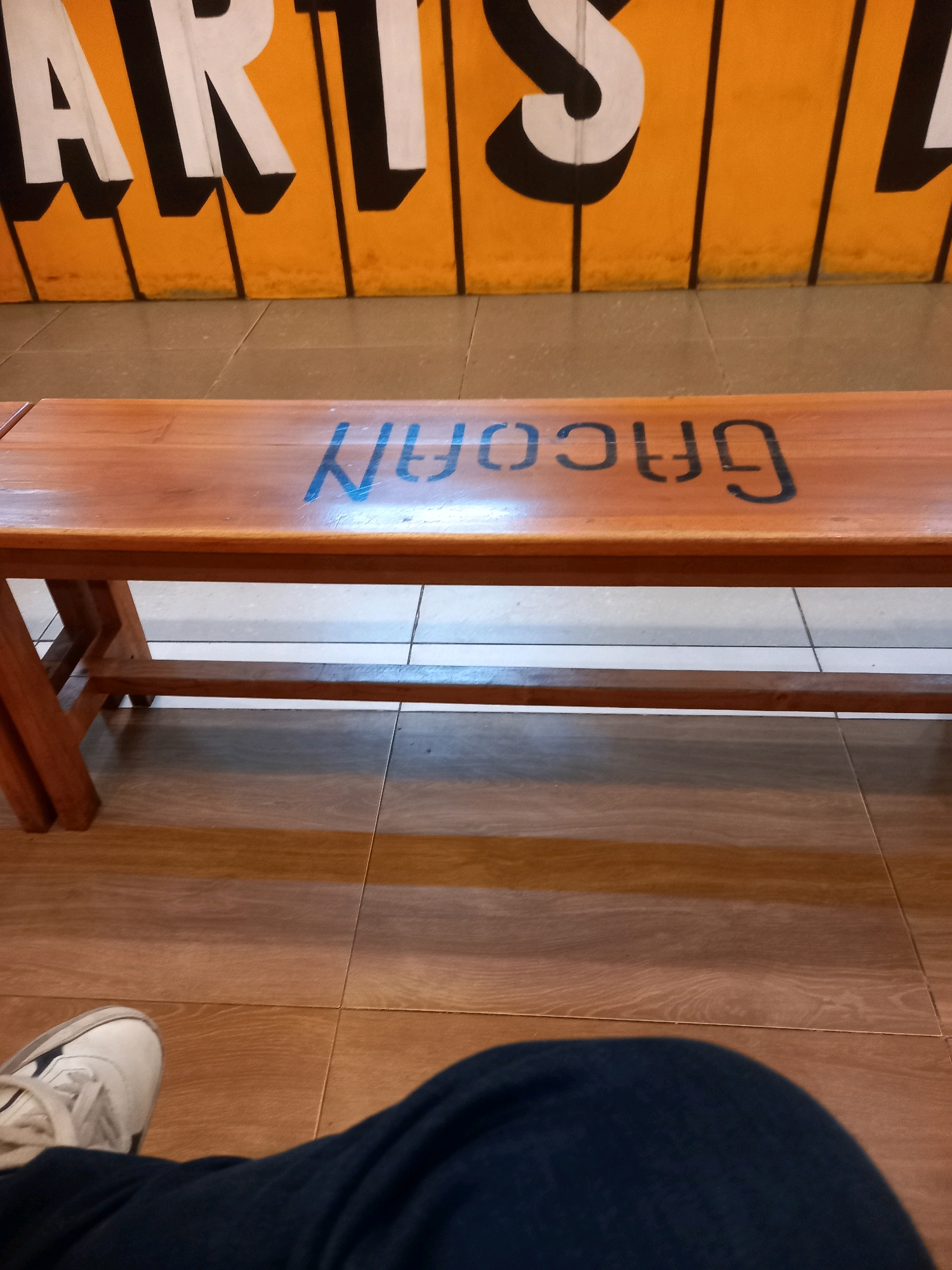 The image shows a wooden bench with blue lettering that appears to spell out "Grocary". The bench is positioned in front of a wall with large orange and white lettering that reads "ARTS". The floor beneath the bench is tiled in a pattern of brown and beige squares. In the foreground, a person's legs, clad in dark blue fabric, and a white sneaker are visible. The setting appears to be an indoor public space, possibly an art gallery or community center, in the city of Pamekasan, Indonesia, given the context provided.