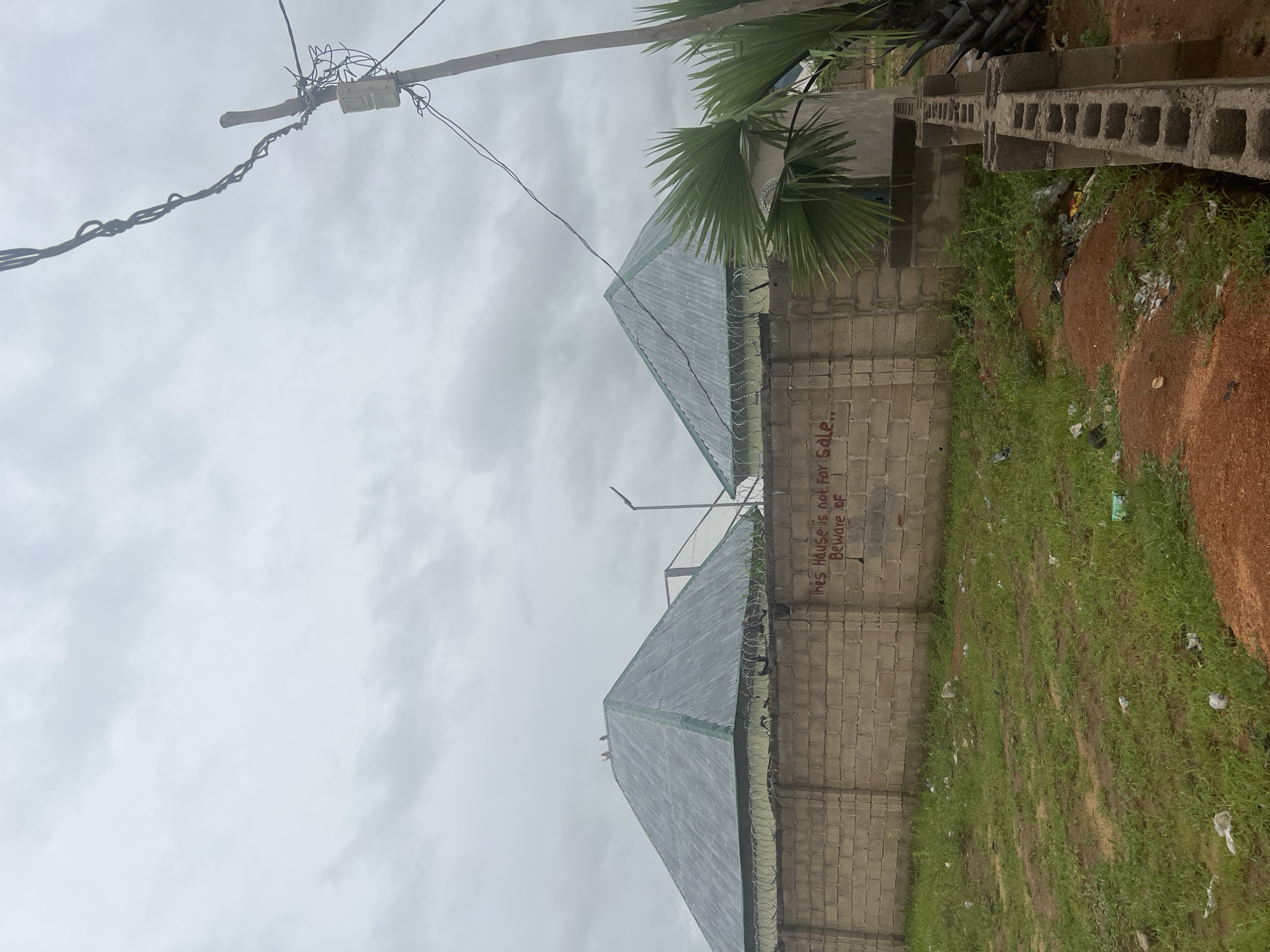 The image depicts a street scene in Howayi, Nigeria, with two conical-roofed houses visible behind a brick wall. The wall is topped with barbed wire, and graffiti in red spray paint reads "This House is not for Sale.. Beware of". The foreground is a patch of overgrown grass with scattered litter. A utility pole with wires stands to the right of the frame, and a palm tree is also visible. The sky is overcast and cloudy. No people are present in the image.