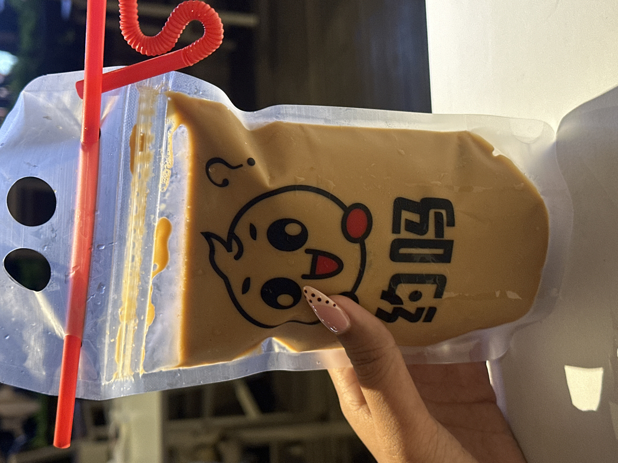 The image shows a close-up of a clear plastic pouch filled with a creamy, tan-colored liquid. Two red, corrugated straws are inserted into the pouch. The pouch has a cartoon face printed on it, featuring large eyes, a smiling mouth, and a red circle on its cheek. To the right of the face, there are two black Chinese characters, which appear to be "奶茶" (nǎichá), meaning "milk tea." A person's hand with manicured nails is holding the pouch. The setting appears to be outdoors, with the background blurred and out of focus, suggesting natural lighting. The overall impression is casual and perhaps related to a beverage purchase.