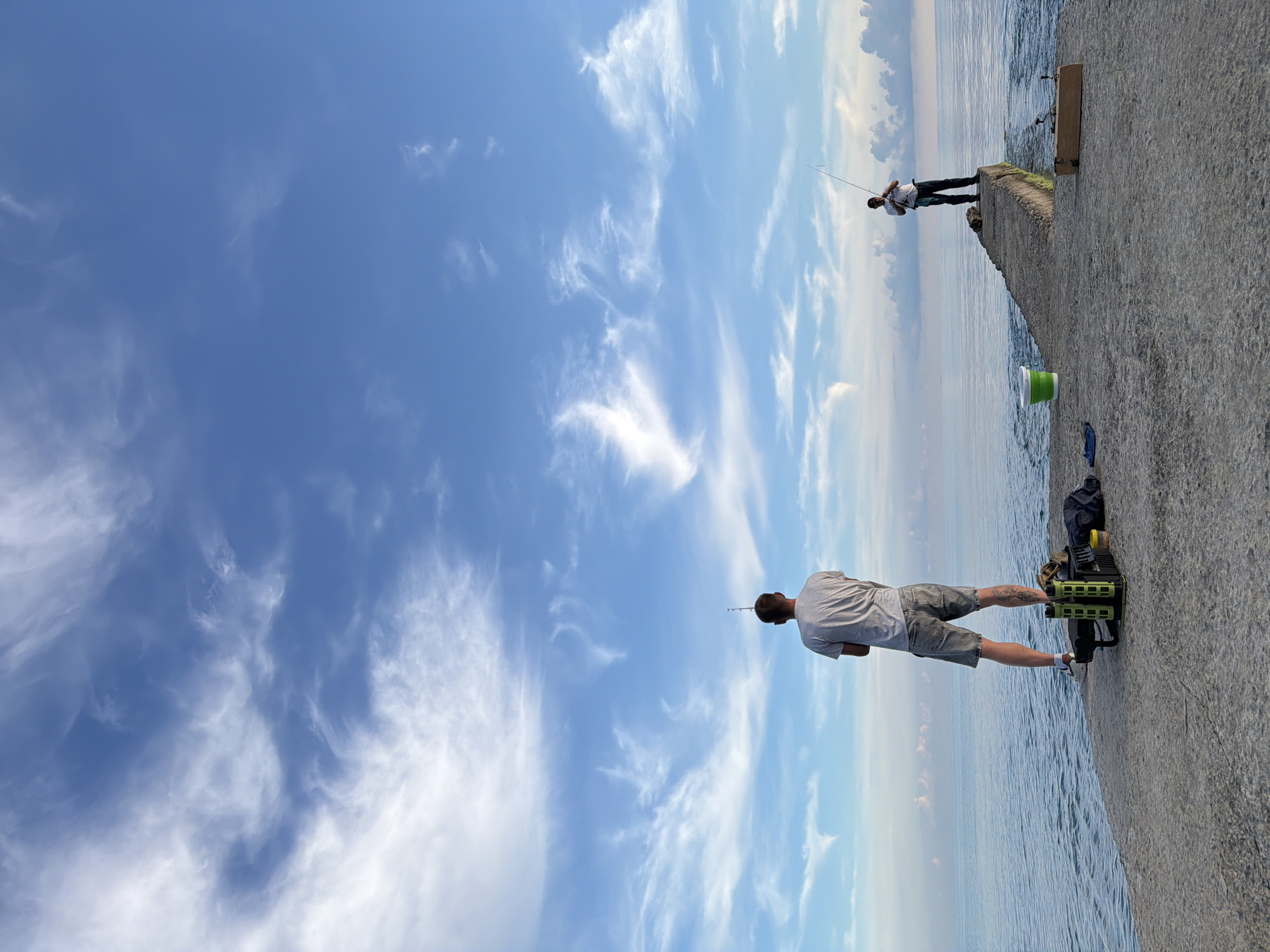 Two men are fishing on a concrete pier extending into the sea under a blue sky with wispy white clouds. The man in the foreground, facing away from the camera, is standing on the wider part of the pier. He is wearing a grey t-shirt and grey shorts, and has a tattoo on his left calf. Next to him are a green fishing rod holder, a black bag, and a yellow container. Further down the pier, the second man, wearing a white t-shirt and dark pants, stands on the narrower section, actively casting his fishing line. A green and white bucket is visible near him. The sea is calm, with a slight ripple on its surface. The sky is a vibrant blue, with soft, elongated clouds scattered across it. The scene appears to be set in Dederkoy, Russia, given the geographical context.