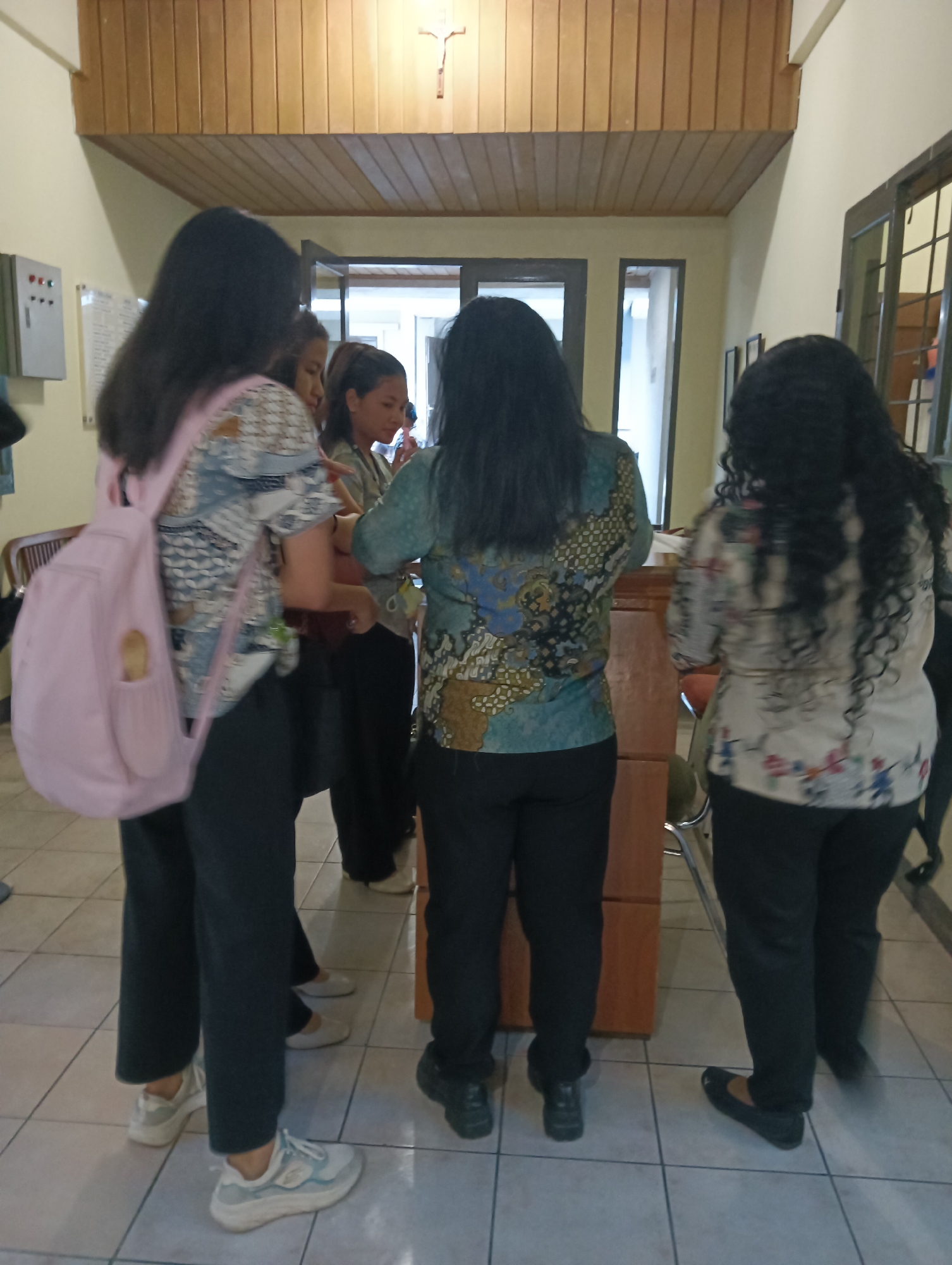 The image depicts an indoor scene, likely within a building in Tomohon, Indonesia, characterized by tiled flooring and wooden paneling on the ceiling. A crucifix is mounted on the wooden ceiling. Several individuals, predominantly women, are visible from behind, dressed in casual attire. One woman in the foreground wears a light pink backpack and a patterned blouse. Another woman to her right wears a teal-colored blouse with an intricate pattern. A third woman, positioned further to the right, is seen in a white blouse adorned with a floral print. The individuals appear to be standing and interacting, though the nature of their interaction is not explicitly clear. In the background, a wooden cabinet or counter is present, and doorways leading to other areas of the building are visible. An electrical panel is mounted on the wall to the left. The overall impression is a casual gathering or waiting area within a possibly institutional or community setting.
