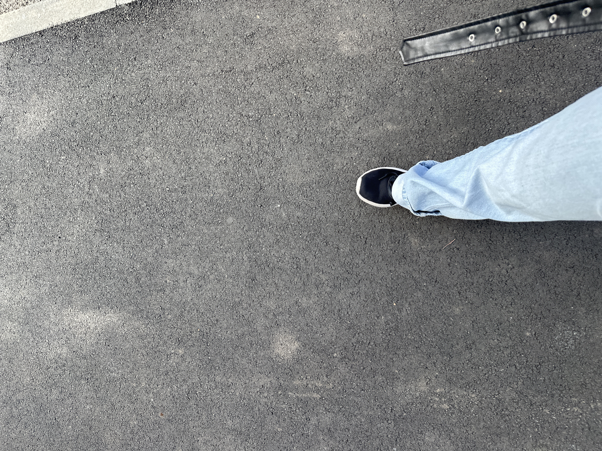 A high-angle, top-down shot captures a person's lower body and the ground beneath them. The subject is wearing light blue jeans, slightly cuffed at the ankle, and a black sneaker with a distinct white sole. Only one foot is visible, positioned towards the lower right of the frame. In the upper right corner, a black strap or belt with several silver eyelets is partially visible. The primary surface is dark gray asphalt, showing a granular texture. A light gray concrete curb or sidewalk edge runs diagonally along the top left side of the frame. The scene is outdoors, likely during daytime, with dry ground. No specific activity beyond standing or walking can be determined. The image provides no specific location cues beyond a generic outdoor setting, and no text is visible. The location context for this image is Kazan, Russia.