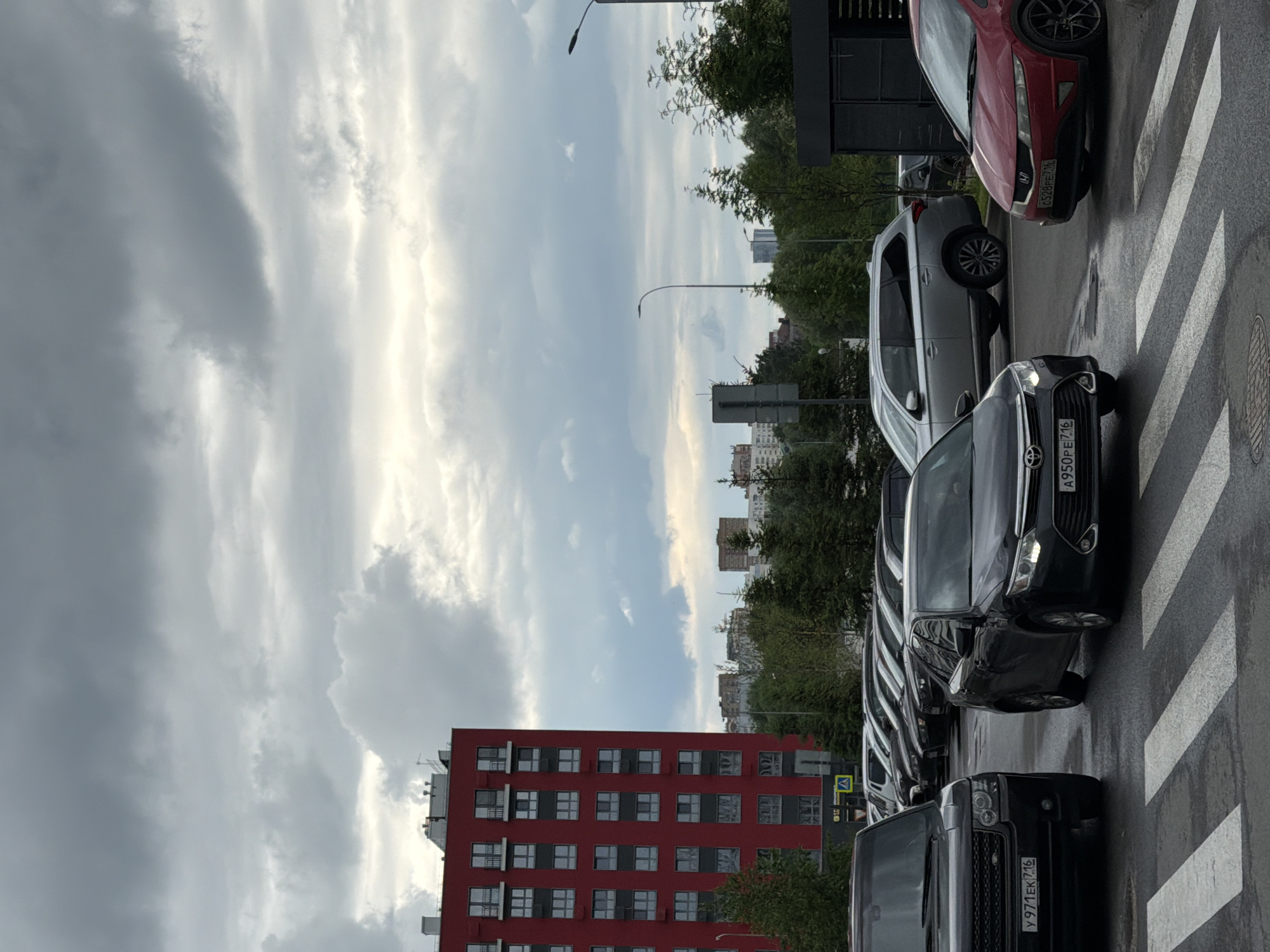 A low-angle exterior shot captures a cloudy day in an urban environment. In the foreground, a black Range Rover and a black sedan are parked at a crosswalk, with a red sedan to the right. The Range Rover has license plate Y 971 EK 716, and the sedan has license plate A950PE 716. In the background, a tall, red apartment building stands to the left, with a row of cars parked in front of it. Further in the distance, more buildings and trees are visible under the overcast sky. The street appears wet, suggesting recent rain.