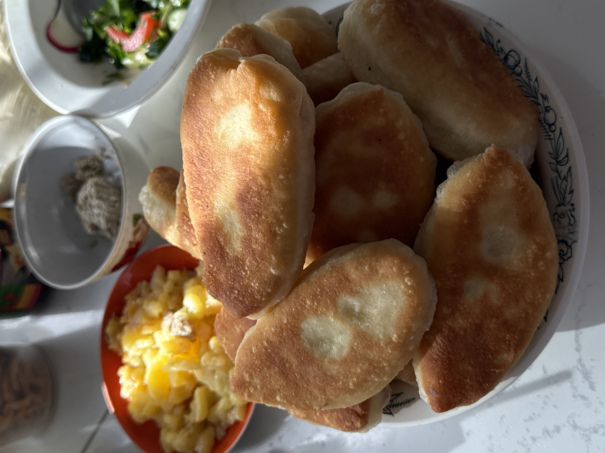 The image depicts a tabletop arrangement of food. A plate piled with fried pastries is the central focus. A bowl of scrambled eggs is positioned to the lower left of the plate. A small bowl containing a beige substance is located towards the upper left. A salad in a white bowl is visible in the upper left corner. The setting appears to be indoors, with a white surface reflecting light. The location is not discernible from the image. There are no people visible.