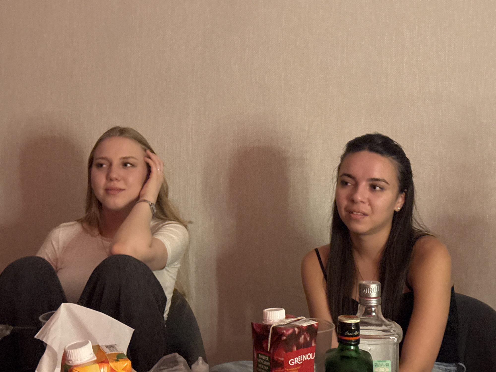 Two young women are seated at a table, with their faces and upper bodies visible. The woman on the left has blonde hair and is wearing a white t-shirt and dark pants. She is looking to her left with her hand on her head. The woman on the right has dark hair and is wearing a black tank top. She is looking to her left, with her hand on her head. In front of them are several bottles and a carton of juice. The background is a textured wall. The image appears to have been taken indoors, in a room that could be in the city of Perm, Russia, based on the objects present.