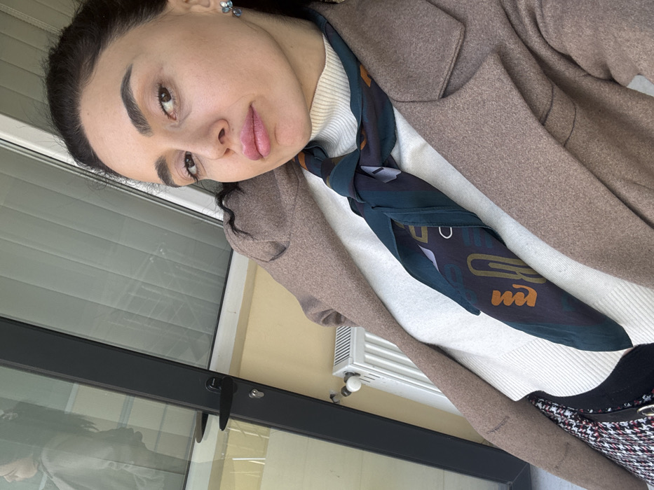 A female subject is depicted in a close-up, self-taken photograph, framing her from the chest up. She has dark hair, defined eyebrows, and full lips, looking slightly towards the camera. She wears a light brown, possibly wool, coat over a cream-colored turtleneck sweater. A dark teal scarf with a pattern featuring stylized shapes, including forms resembling paper clips, is tied around her neck. A black and white patterned fabric, possibly a skirt, is visible on the lower left, along with a portion of a black bag featuring a silver buckle. The background on the left includes a dark-framed glass door or window reflecting a blurry, light-colored figure. Below this, a white multi-finned radiator is mounted against a pale yellow wall. To the subject's right, a window with horizontal white blinds is visible. The setting appears to be an interior space or enclosed balcony of a building. The image contains no specific geographical markers identifying a precise location within Tbilisi, Georgia.