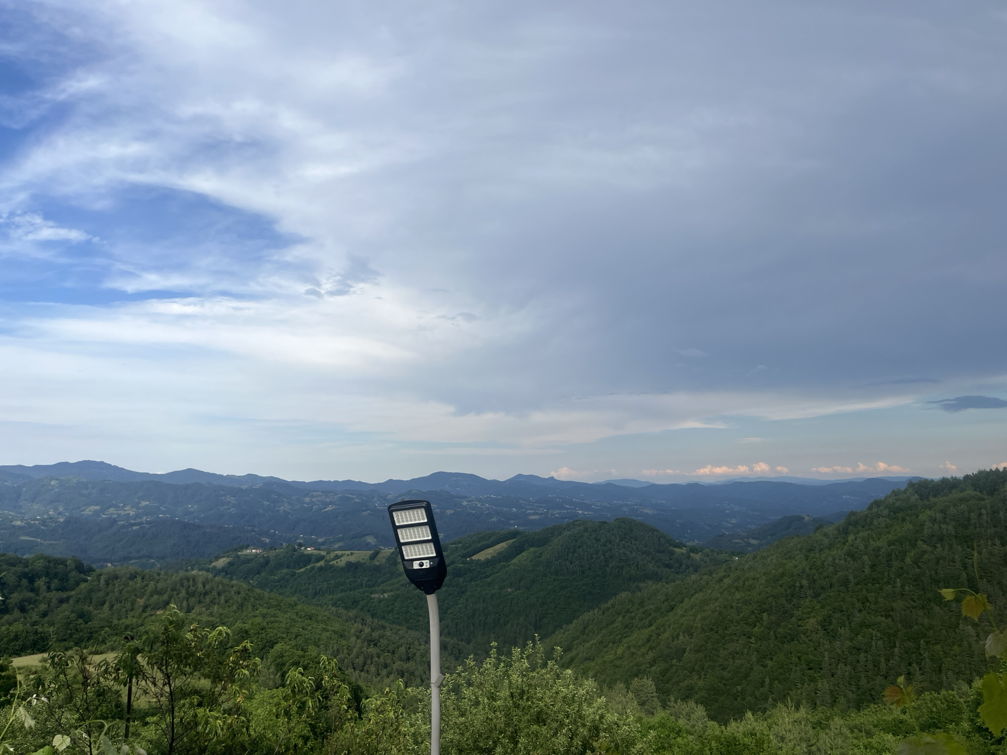 A black solar-powered LED street light stands on a pole in the foreground, angled slightly to the right. It is positioned against a backdrop of rolling green hills and mountains stretching into the distance under a partly cloudy sky. The hills are covered in dense green trees, with some scattered houses visible on the slopes further back. The sky is a mix of blue and grey clouds, with brighter patches of blue peeking through. The overall scene depicts a natural landscape with a modern utility structure. The location is the city of Startsevo, Bulgaria.