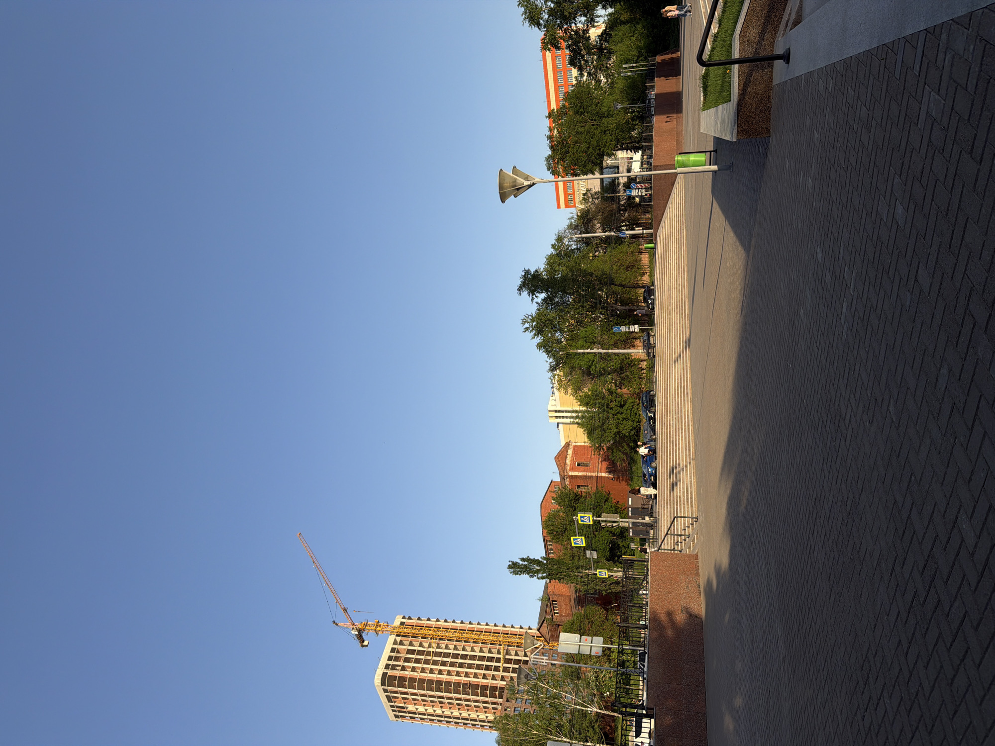 The image depicts an urban scene in Izhevsk, Russia, with a clear blue sky dominating the upper portion. A tall building under construction is visible in the lower left, featuring a yellow construction crane. A street with pedestrian crossings and street signs is lined with trees and various buildings, including red brick structures. A paved walkway extends along the right side of the frame, composed of interlocking bricks. Some people are visible walking in the background. The scene is bathed in sunlight, casting shadows on the walkway and buildings.