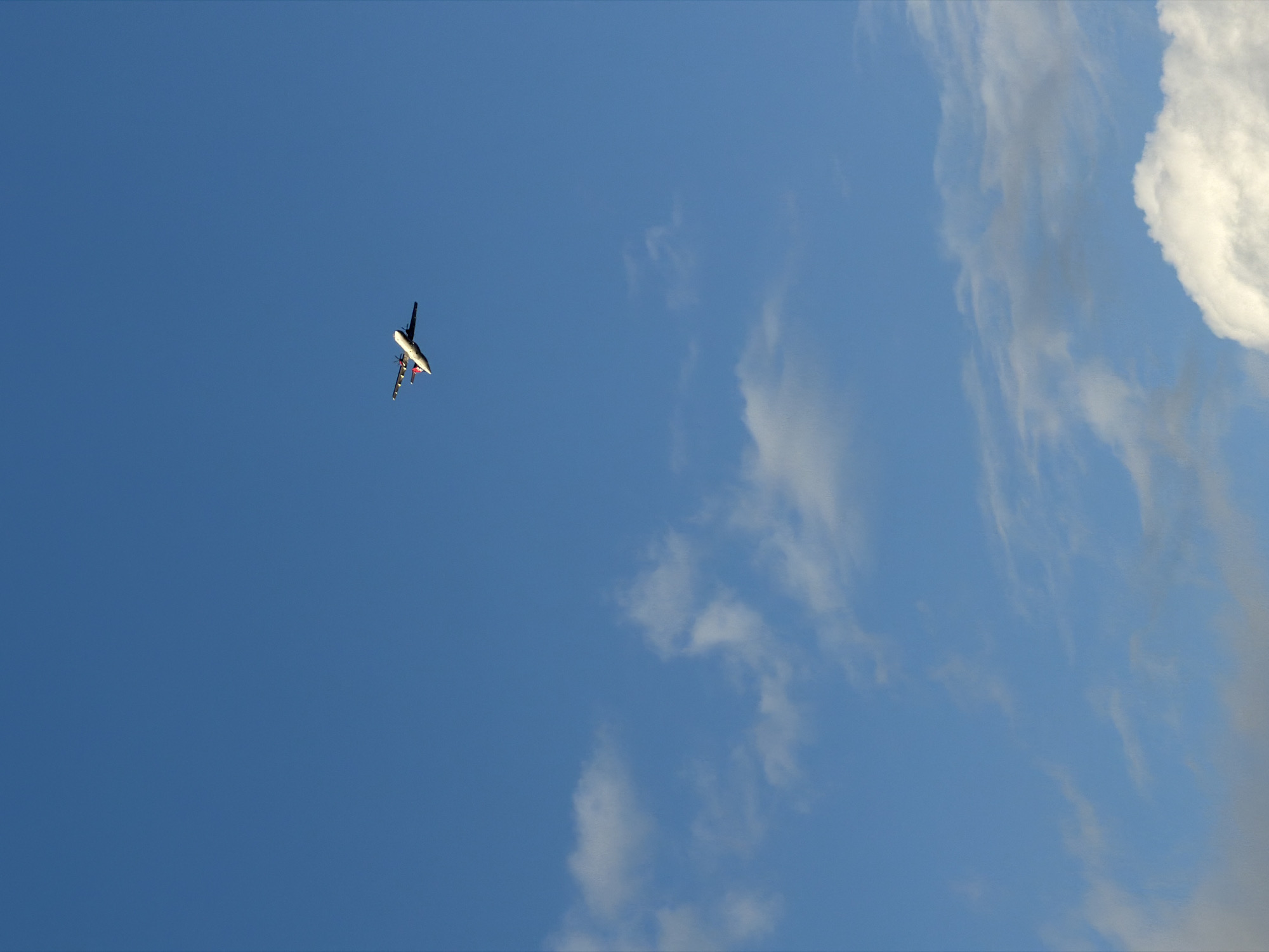 The image shows an airplane in flight against a blue sky with scattered clouds. The airplane is in the upper left quadrant of the image. A large, puffy cloud is visible in the upper right corner, with wisps of other clouds in the center and bottom right. The location is Gošići, Montenegro.