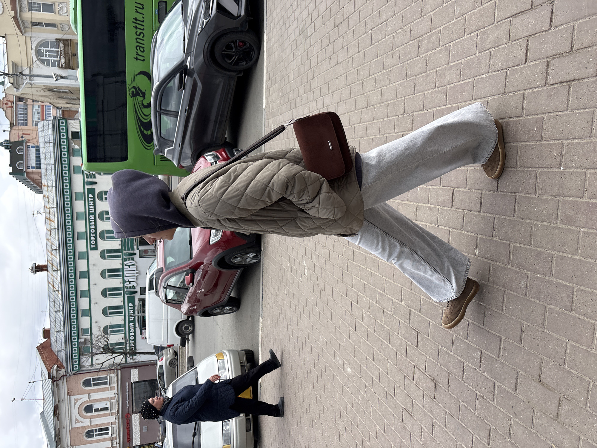 A person wearing a gray hooded sweatshirt, a light brown quilted jacket, light gray wide-leg jeans, and brown shoes is walking on a brick sidewalk. The person has a dark brown shoulder bag. In the background, there are various cars, including a green bus with "transitlt.ru" written on it, a gray SUV, a red car, a white van, and a white car with a person standing next to it. The buildings in the background have signs in Russian, including "ТОРГОВЫЙ ЦЕНТР", placing the location as Ulyanovsk, Russia. The sky is overcast. Another person, wearing a black jacket and a black hat, is walking away from the camera next to a white car.