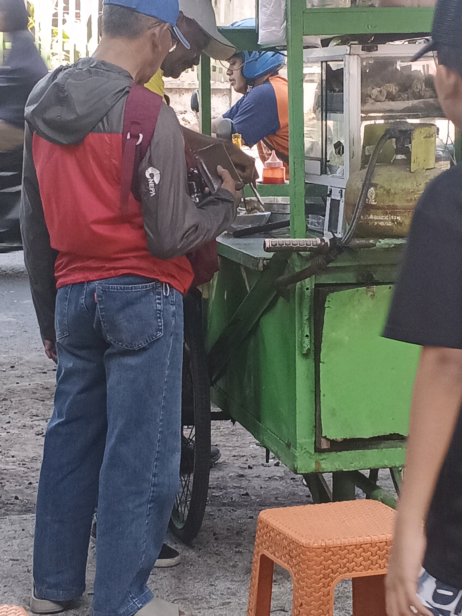In Krian, Indonesia, a group of individuals gather around a food cart. The cart, painted a vibrant green, is laden with various items, including a prominent yellow gas cylinder and glass display cases filled with food. Two men in the foreground are the primary focus. One man, wearing a red and grey jacket and blue jeans, is holding a small object. Another man, partially visible, stands to his right. Behind them, a person in an orange and blue uniform appears to be preparing food. The environment suggests a street-side setting with a paved ground and natural light.