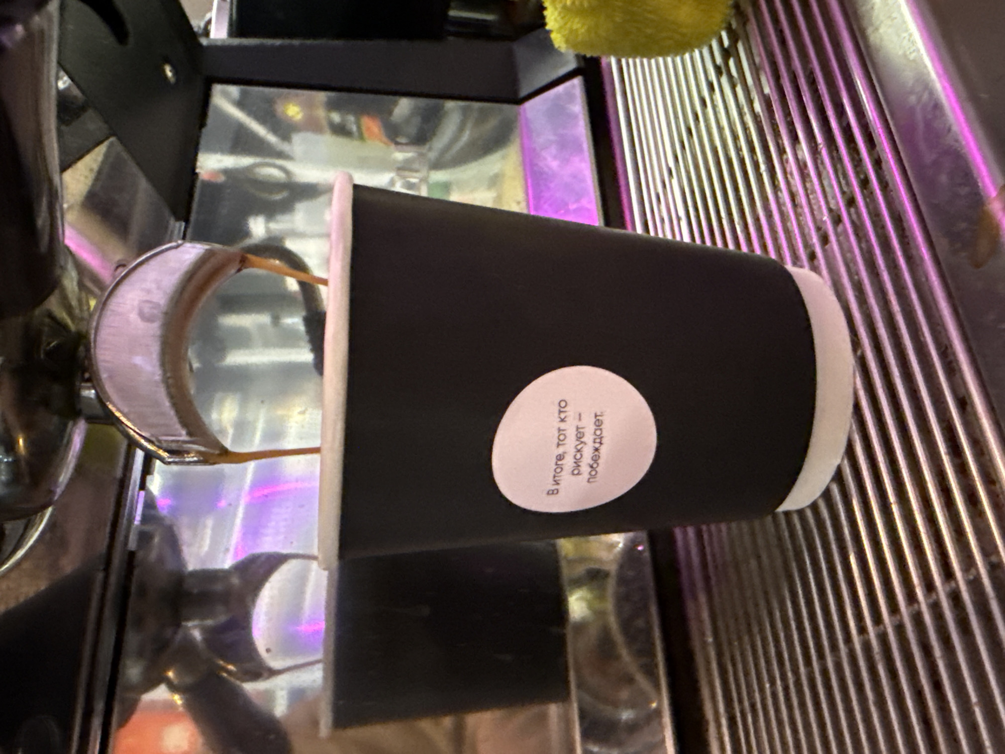 A black disposable coffee cup is positioned under the spouts of an espresso machine, receiving a thin stream of what appears to be coffee or espresso. The cup has a white rim. The scene is set on the metal drip tray of the espresso machine, which features a ribbed grate and reflective surfaces. A yellow cloth or sponge is partially visible in the upper right background. Bright purple and pink light illuminates parts of the metallic surface, suggesting neon or LED lighting in the environment, possibly a coffee shop or cafe.
A circular white sticker is affixed to the side of the black coffee cup, displaying text in Cyrillic. The text reads: "В итоге, тот кто рискует — побеждает." This translates to: "In the end, he who risks — wins."
No people are visible in the image. The overall impression is an active coffee preparation station, likely in Moscow, Russia, given the provided context. The metal grate on the drip tray shows some signs of residue or use.
