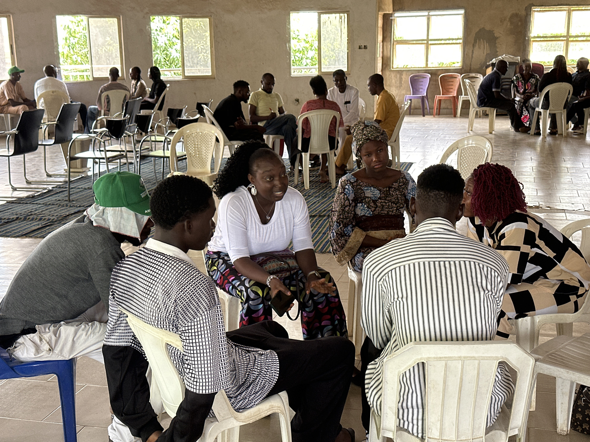 This image depicts an indoor gathering of people in what appears to be a community hall or a large room in Sabon Tasha, Nigeria. The setting is functional, with a tiled floor and large windows that allow natural light to filter in, suggesting it is daytime. The atmosphere seems relaxed and conversational.
The primary subjects are several individuals, men and women, seated in plastic chairs arranged in small groups, suggesting a meeting or social gathering. Some people are facing each other, engaged in conversation, while others are looking towards the camera or in other directions. Their attire is casual, with some wearing traditional African clothing and others in more contemporary outfits.
One woman in the foreground is wearing a white t-shirt and a vibrantly patterned skirt, holding a mobile phone in her hand. Another woman next to her has red curly hair and is wearing a patterned outfit. In the mid-ground, a group of men and women are seated, some with notebooks and pens, possibly indicating a discussion, workshop, or educational session. To the left, a man wearing a green cap is seated, facing away from the camera.
The overall impression is one of a community event or meeting taking place in a communal space. The lighting suggests it is daytime, and the presence of natural light from the windows indicates fair weather. There are no visible signs of distress or urgency, implying a calm and perhaps productive environment. The image captures a moment of social interaction and potential community engagement.