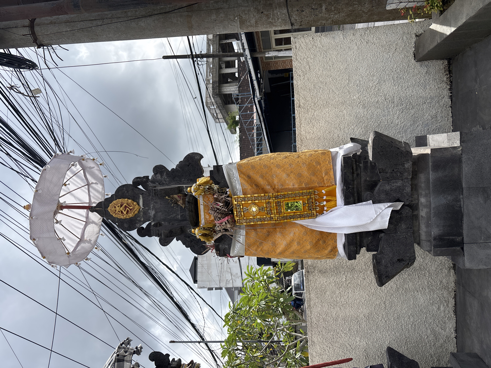 In Kuta Selatan, Indonesia, a stone shrine is adorned with a white umbrella and vibrant yellow and white fabric. The shrine is situated outdoors with a textured, light-colored wall behind it and a dark paved ground in front. A complex network of overhead electrical wires crisscrosses the cloudy sky. In the background, a white van is visible, along with several buildings, one of which appears to be under construction. A small plant with red flowers is growing to the right of the scene. The image captures a street-side view of the shrine, which is a common sight in Balinese towns.