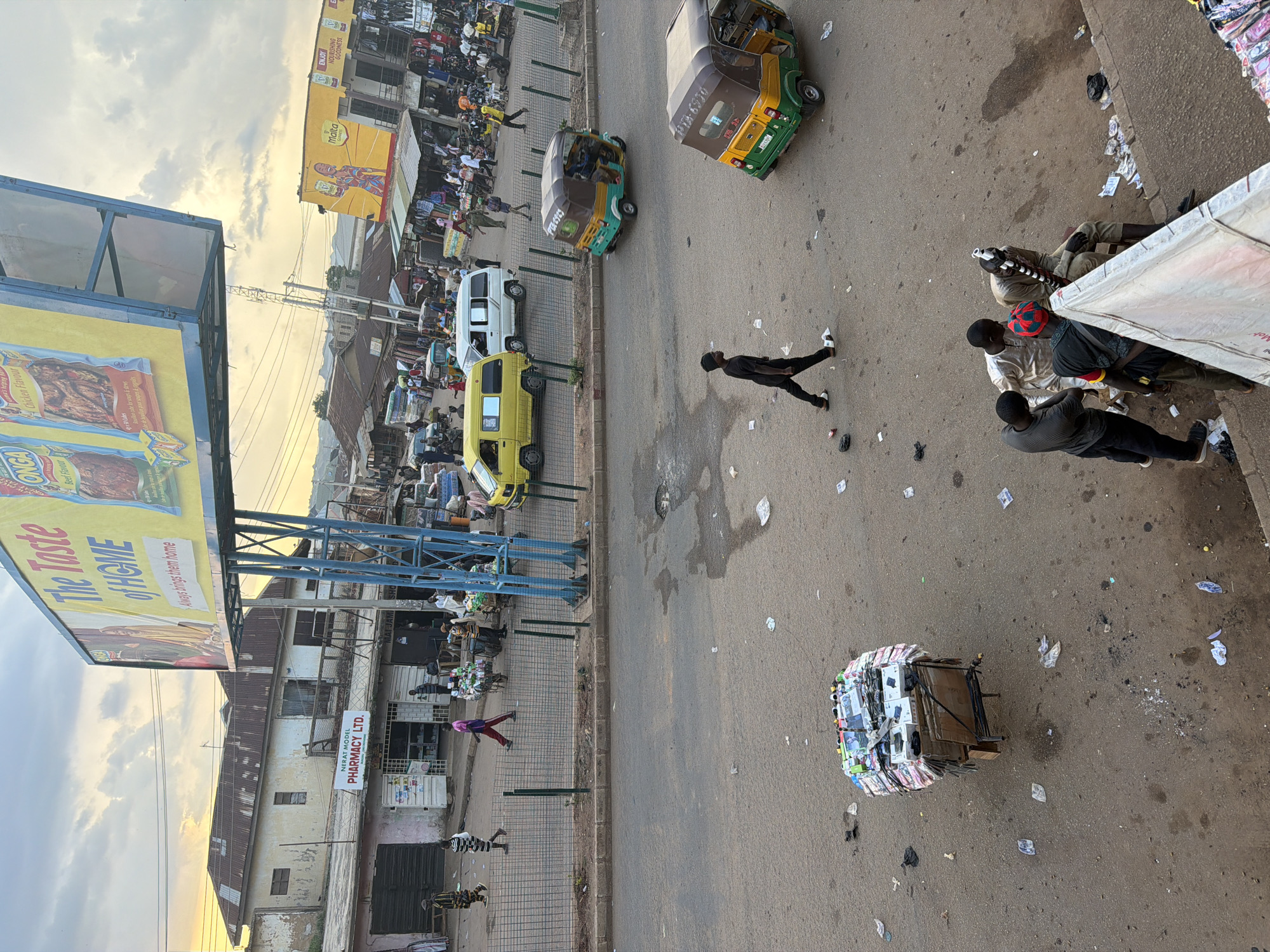 This is a high-angle, vertical photograph taken in Bukuru, Nigeria, showing a bustling street scene. The sky is overcast with a sunset glow, suggesting the time of day is late afternoon or early evening. The street is paved and littered with various pieces of paper and debris.
In the foreground, several people are visible. A man in a black shirt and pants walks across the street. Further down, a person in a purple outfit walks past a fenced area. Nearer the bottom of the frame, two individuals in striped clothing are standing. In the mid-ground, a group of people are gathered, some sitting under an awning.
Various objects populate the scene, including several vehicles. A yellow minibus and a white van are parked along the roadside. Two three-wheeled auto-rickshaws are also present, one parked and another in motion. A prominent object in the foreground is a large billboard advertising "The Taste of HOME" and "Chicken Flavour" and "Beef Flavour" food products. Another billboard further in the background advertises "Malta" and "ENJOY NOURISHING GOODNESS." A smaller sign reads "NERAT MODEL PHARMACY LTD." A peculiar sight is a large, circular pile of what appears to be stacked goods, possibly books or packaged items, on a trolley in the middle of the road.
The overall impression is one of a busy marketplace or urban street with significant commercial activity and pedestrian traffic. The setting appears to be a somewhat cluttered but vibrant commercial area in Nigeria.