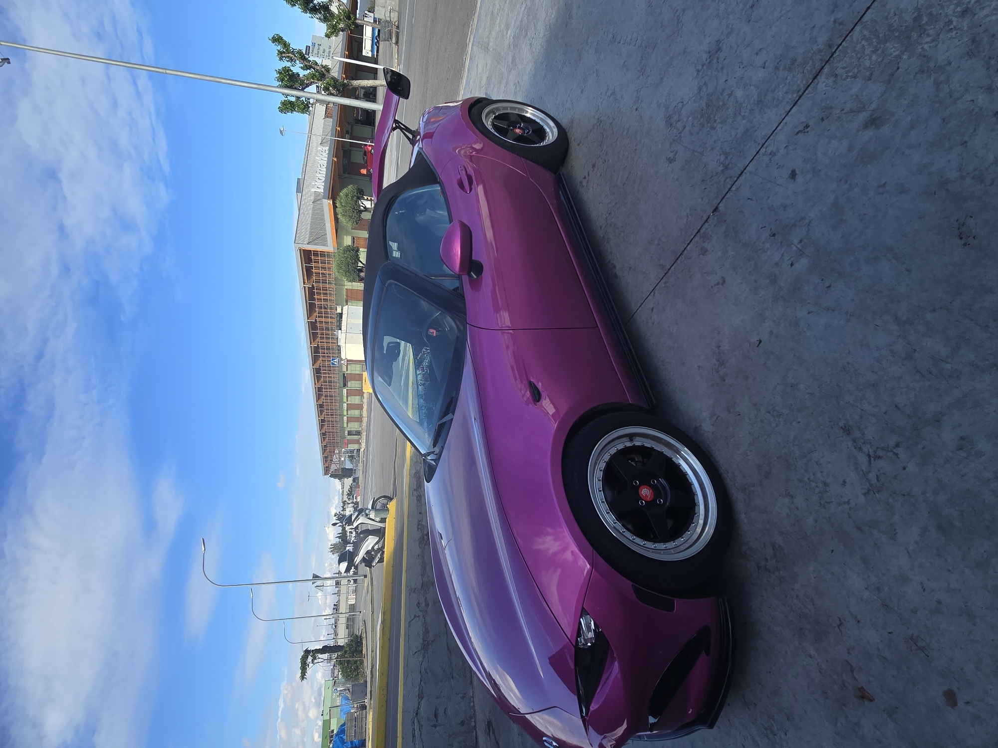 The image shows a pink convertible car parked on a concrete surface. The car has black and silver rims, a black roof, and a rear spoiler. In the background, there is a building with the word "Diagonal Mar" visible. There are streetlights, palm trees, and other buildings in the distance. Motorbikes are parked beside the street. The sky is blue with some clouds. Based on the "Diagonal Mar" sign, the location is likely in the Diagonal Mar area of Barcelona, Spain.