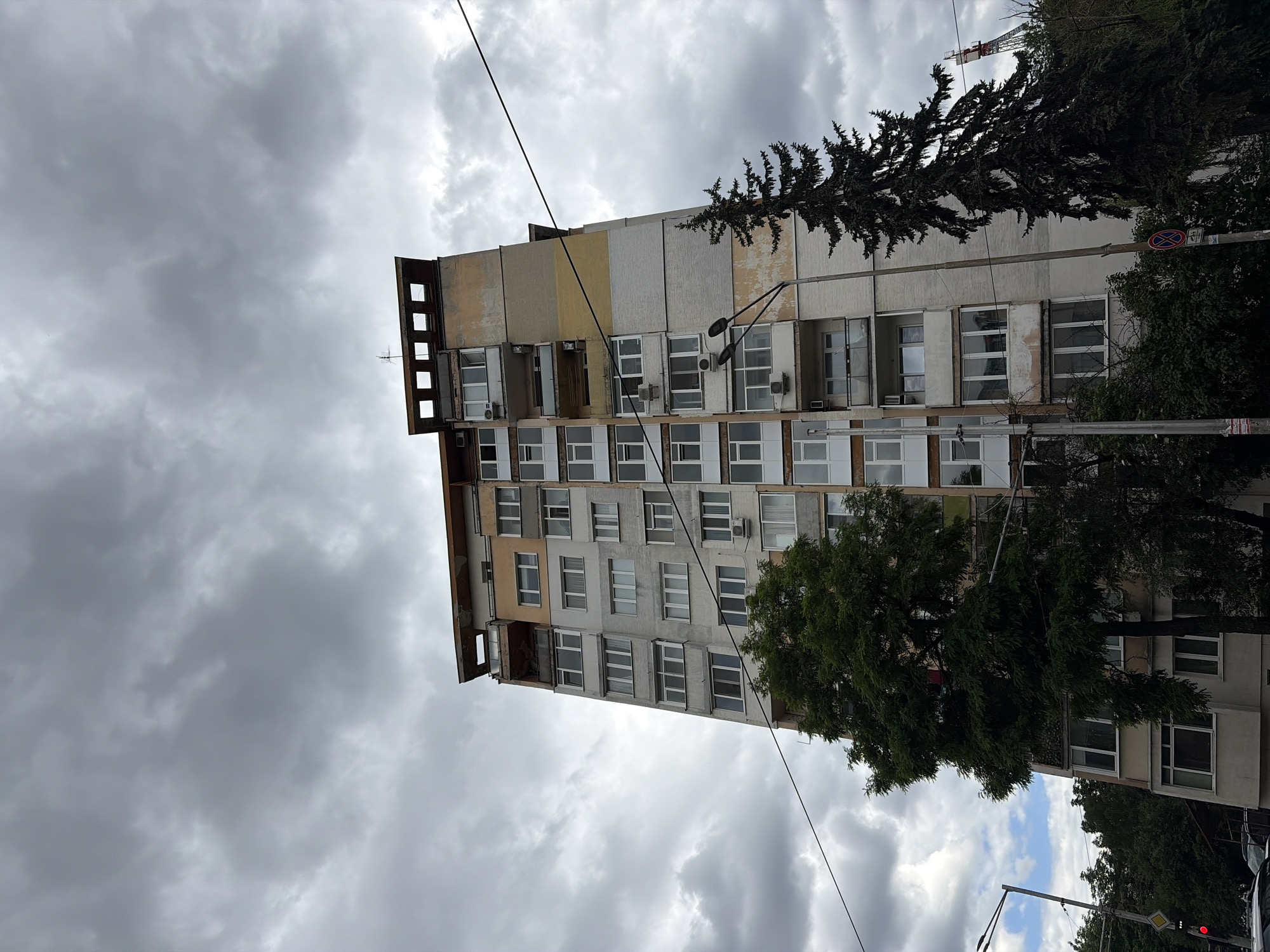 A tall, multi-story apartment building with a weathered facade of beige, light gray, and yellow panels dominates the center of the frame, set against a cloudy, overcast sky. The building features numerous windows and balconies. A large, dense green tree is positioned in front of the lower section of the building on the left side. To the right of the building, a tall, dark evergreen tree stands partially obscuring the structure. A streetlamp with two lamps is visible on the right side of the building, and power lines crisscross the sky. In the background, the upper portion of a tall, lattice-like structure, possibly a crane or tower, is visible. The ground level shows glimpses of other trees and what appears to be part of a street or parking area with a traffic signal.