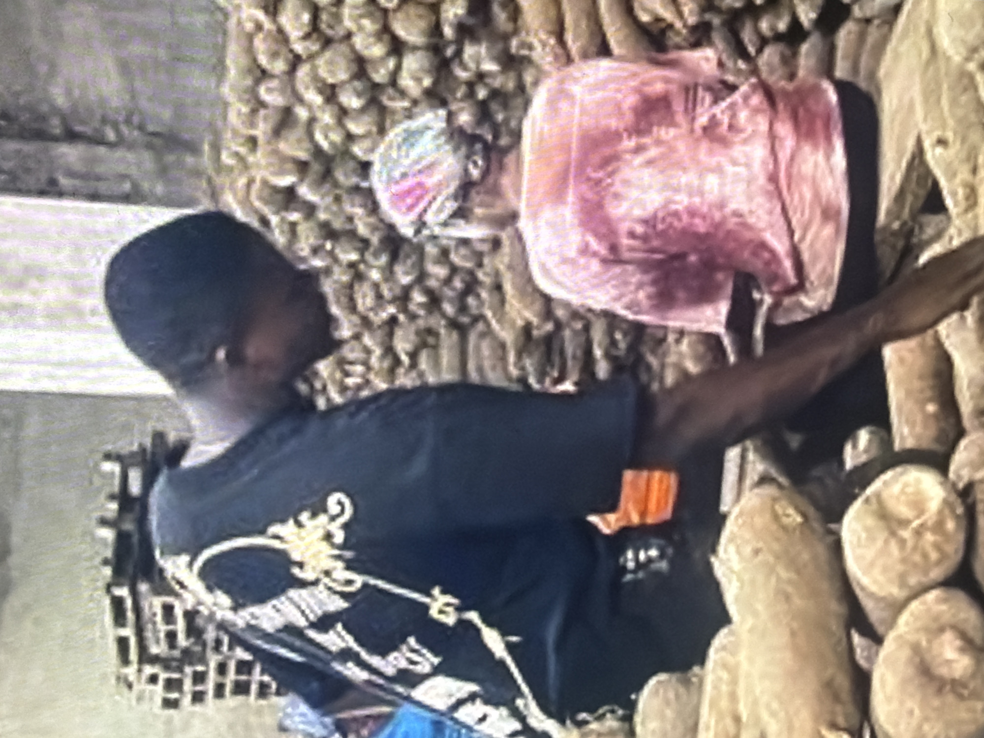 In Bukuru, Nigeria, two individuals are seen amidst a large collection of yams. A man wearing a dark t-shirt with gold embellishments on the back is bent over, interacting with a woman who is seated. The woman is wearing a pink shirt and a headscarf with colorful patterns. The yams are stacked in large quantities, filling much of the background and foreground of the image. Pallets are also visible in the background, suggesting a storage or distribution area. The overall environment appears to be an outdoor or semi-outdoor space, likely a market or storage facility for agricultural produce.