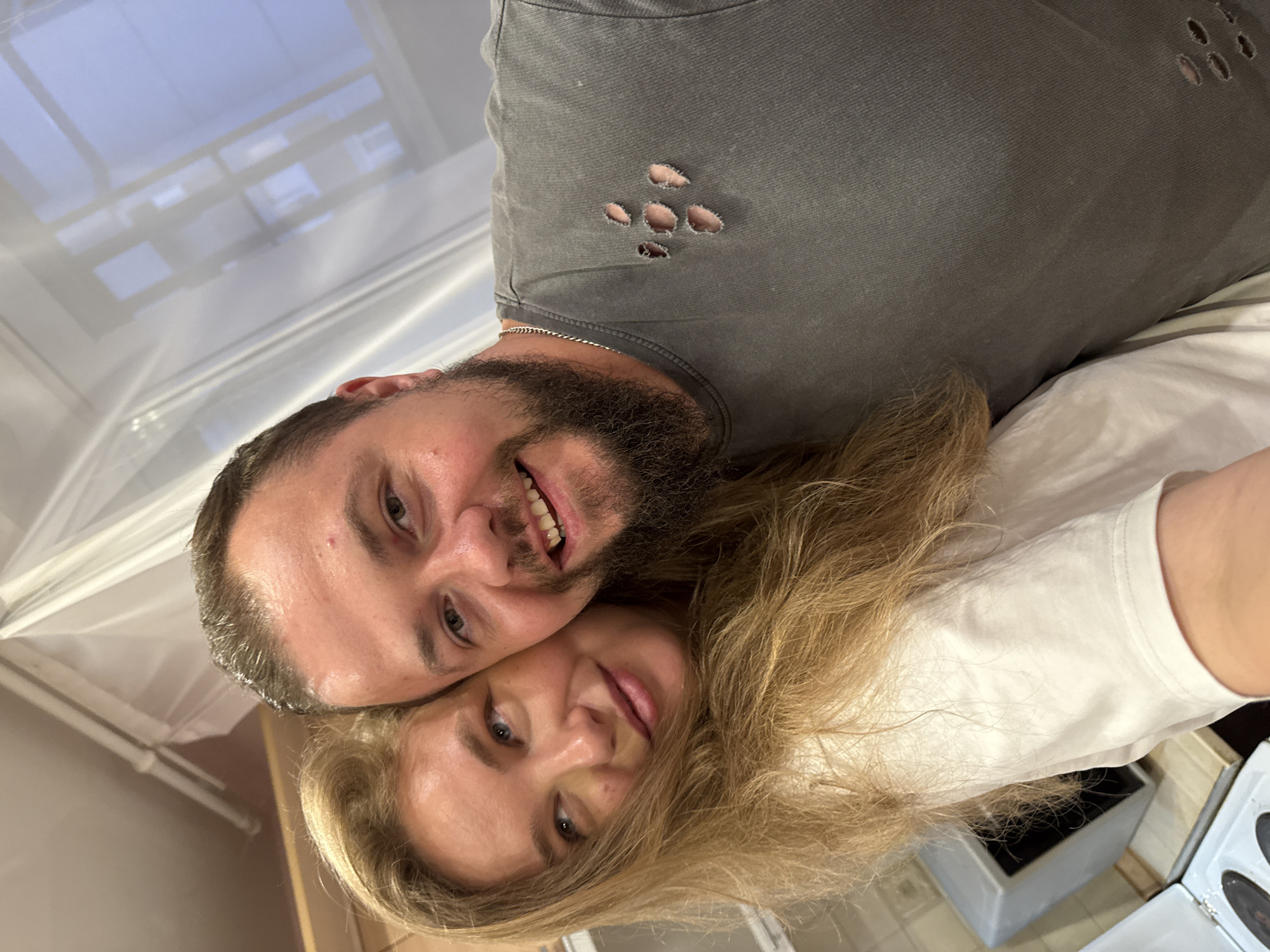 The image shows a close-up selfie of a man and a woman. The woman, on the left, has long blonde hair and is wearing a white t-shirt. She is leaning her head on the man's shoulder. The man, on the right, has short brown hair, a beard, and is wearing a grey t-shirt with several rips on the chest. Both are smiling and looking towards the camera. The background appears to be an indoor setting, possibly a kitchen, with a stove and a microwave visible. The lighting is artificial and warm. The overall scene suggests a casual and affectionate moment between the couple. The location is identified as Vladimir, Russia.