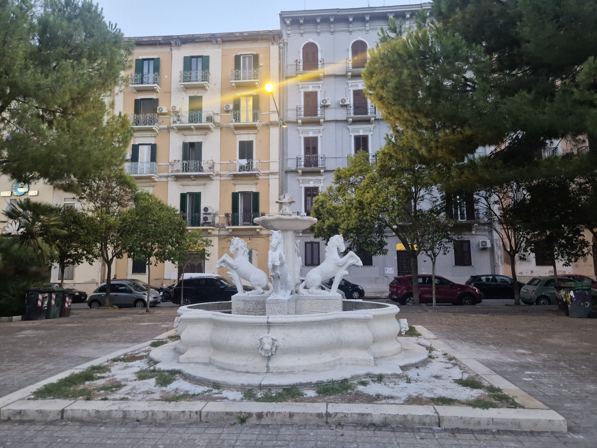 The image shows a public fountain with horse sculptures in the foreground, in front of two multi-story buildings. The buildings are light yellow and light gray, with balconies and green shutters. There is a street with cars parked along it. Trees are visible on the left and right sides of the image. The fountain is white and appears to be made of stone, with three horse sculptures and smaller decorative elements. The ground in front of the fountain is paved with cobblestones and has patches of dirt and grass. The sky is overcast. The scene appears to be in Taranto, Italy.