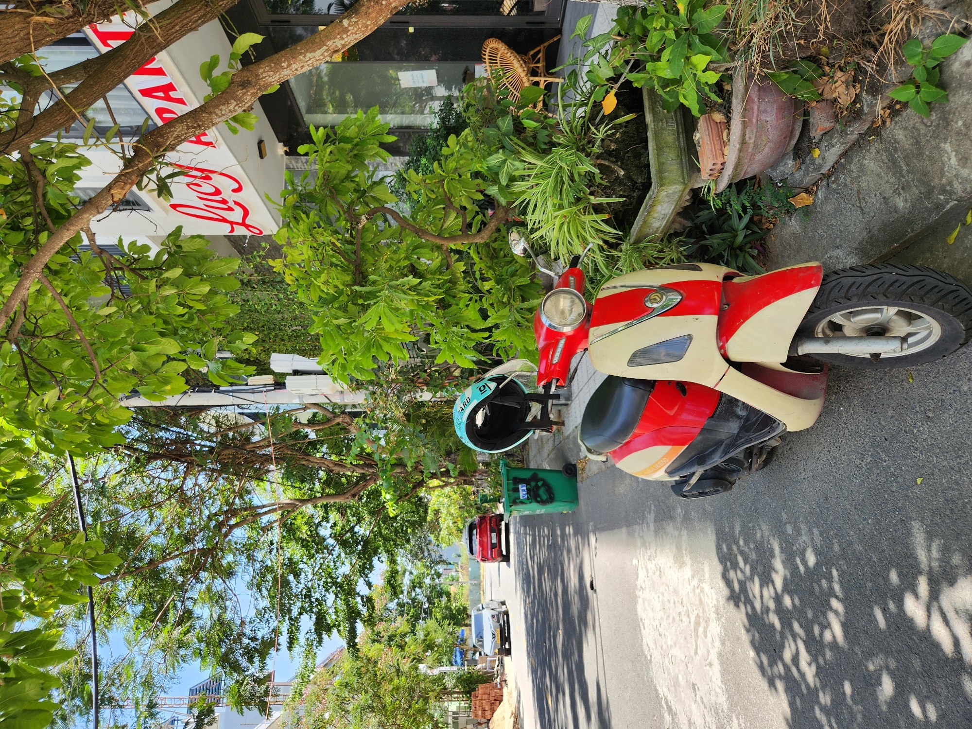 The image captures a street scene in Da Nang, Vietnam, on a sunny day. A red and cream colored scooter is parked on the side of a paved road, next to a sidewalk with lush greenery. A teal helmet with "LEOPARD" written on it rests on a black stand behind the scooter. In the background, a multi-story building with the name "Lucy Apart" visible in red script is partially obscured by trees. Further down the street, two cars are driving, and a green trash bin is situated near the sidewalk. The environment suggests a residential or commercial area, with buildings and trees lining the street.