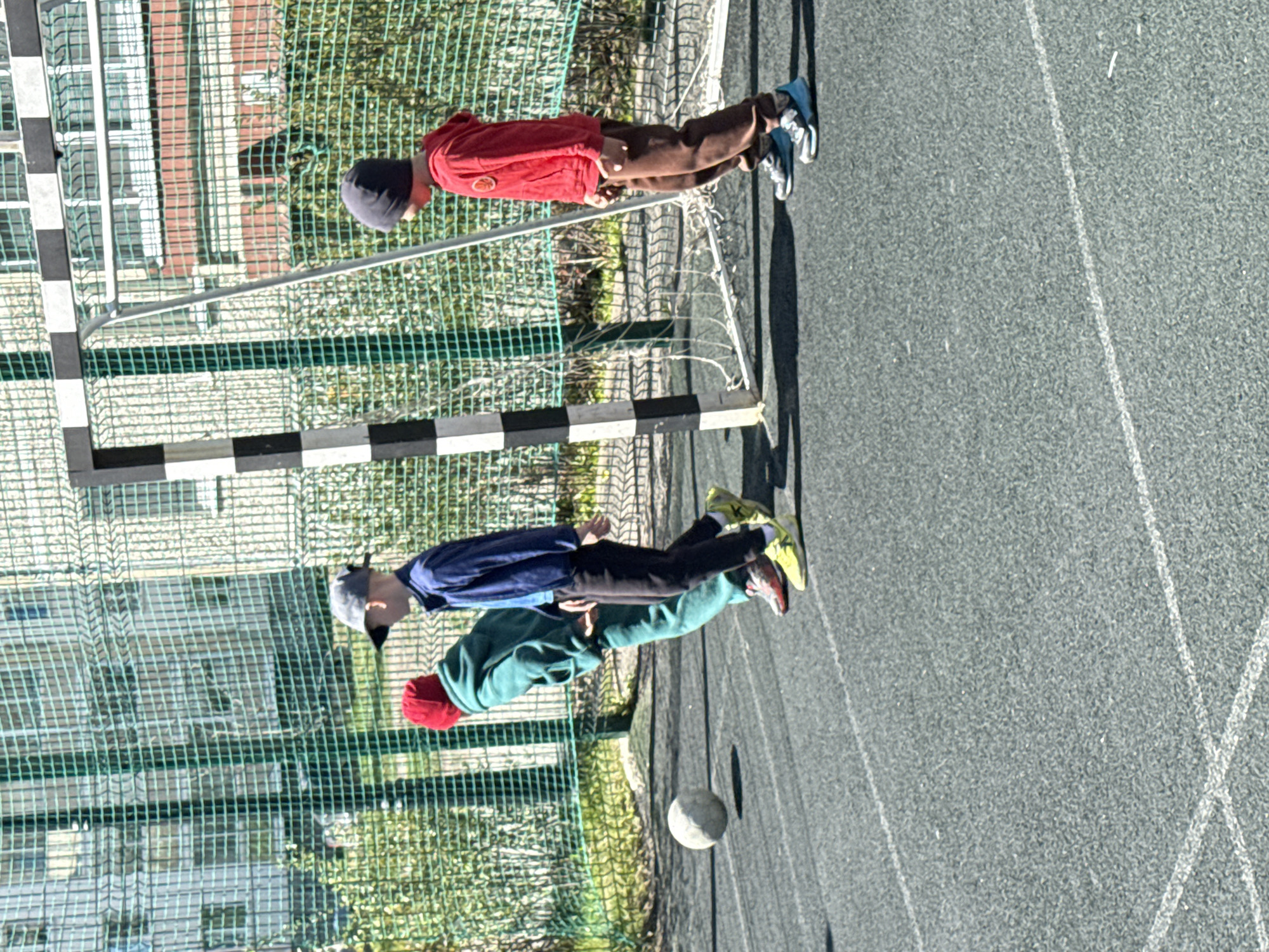 Three boys play on a soccer field in Yekaterinburg, Russia. Two are near a soccer goal and a ball. The other is by a soccer goal further away. The surface is asphalt with white lines. Behind the fence are buildings and vegetation.