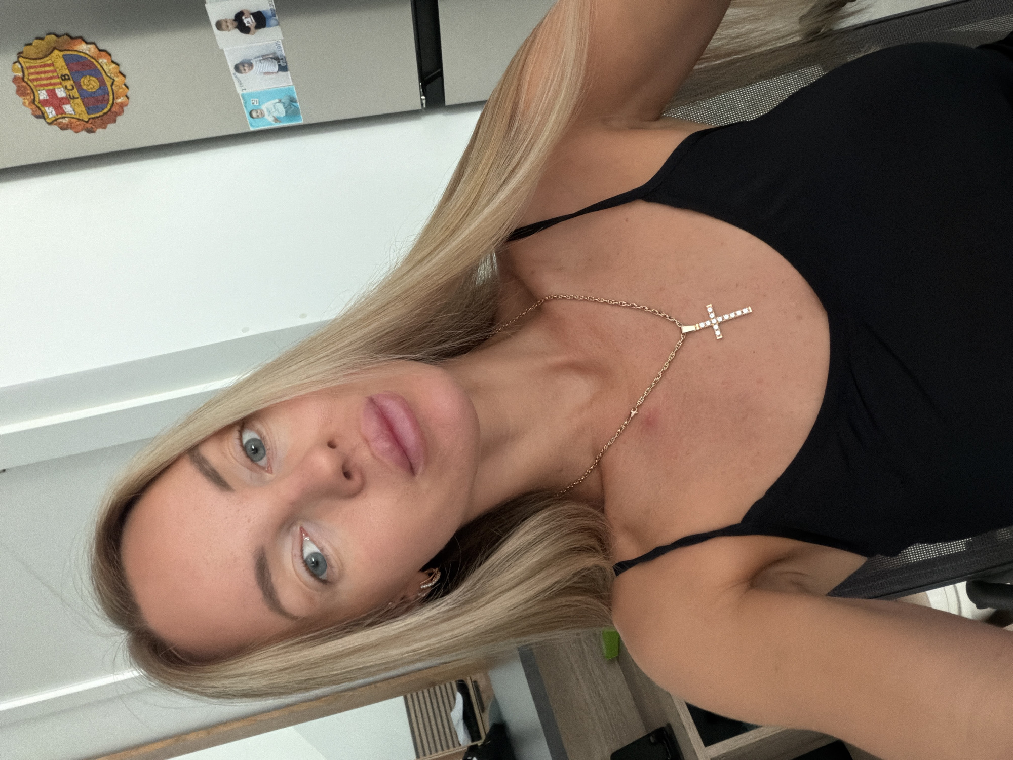 An outdoor, eye-level, close-up selfie shows a blonde woman with blue eyes and full lips. She is wearing a black tank top and a gold chain with a jeweled cross pendant. Her blonde hair is long and straight. In the background, a white wall and a gray refrigerator are visible. The refrigerator has a FC Barcelona crest sticker and several small photos attached. A wooden shelf unit is partially visible to the left of the woman. The overall impression is that the photo was taken inside a home.