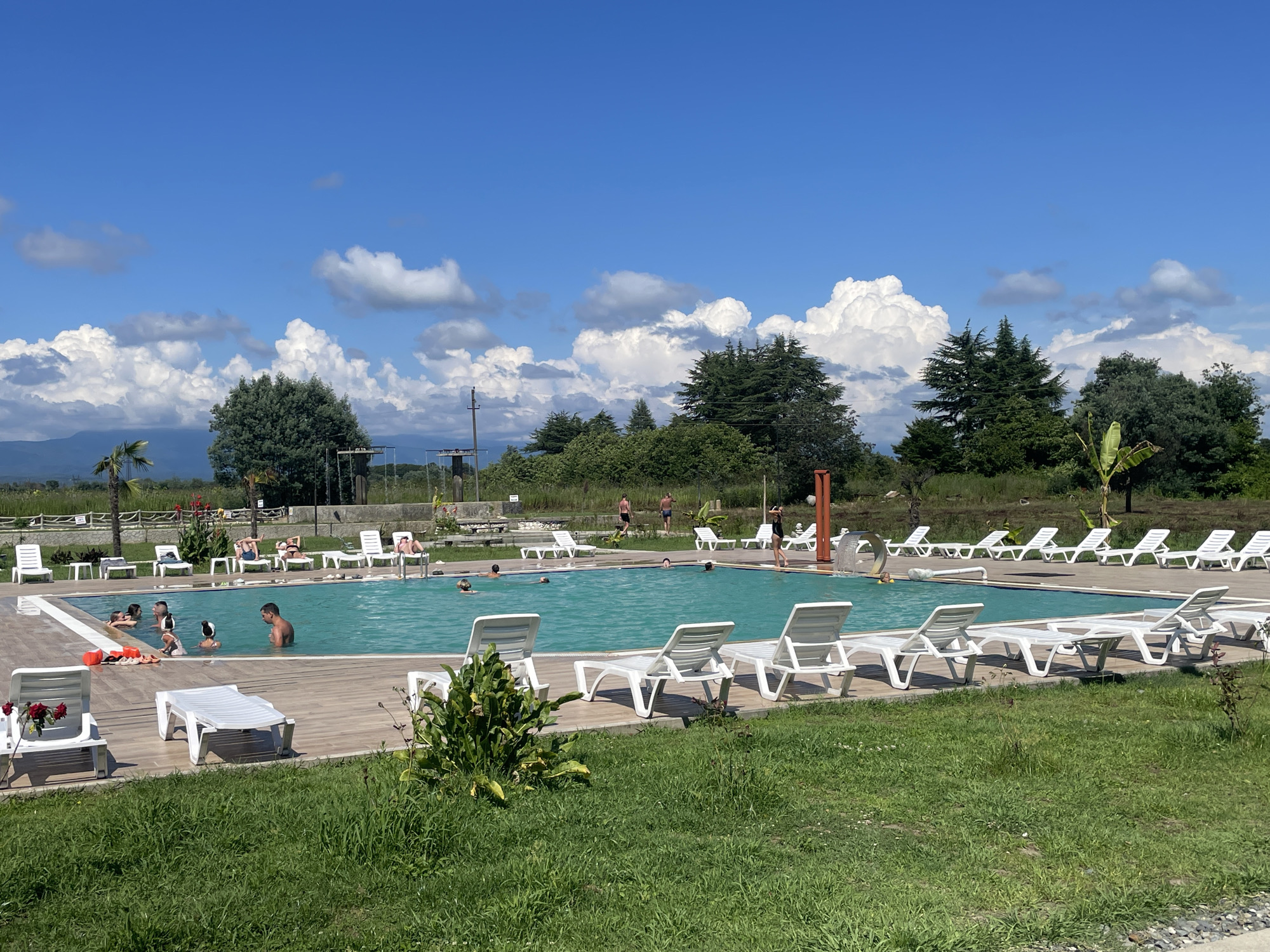 A sunny day at a swimming pool resort in Adzyubzha, Abkhazia. The pool is filled with clear blue water, and several people are swimming or relaxing on lounge chairs. The pool area is surrounded by a wooden deck, and there is a lush green lawn in the foreground. In the background, there are trees and a clear blue sky with white clouds. The scene appears peaceful and idyllic, suggesting a vacation destination.