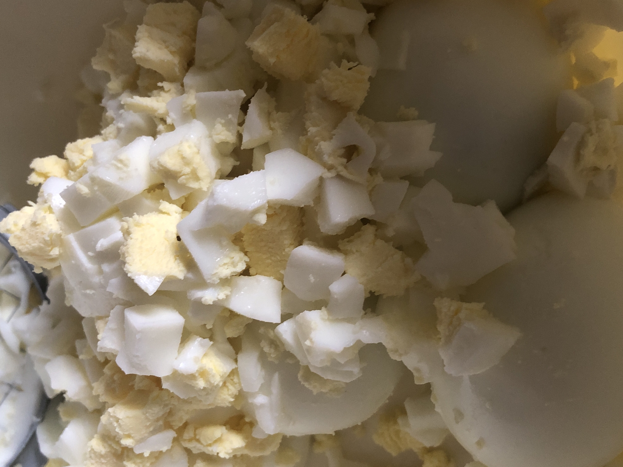 A close-up shot shows a bowl of chopped hard-boiled eggs. The eggs are cut into small, irregular pieces, with both the white and yolk visible. Some pieces are larger and more defined, while others are crumbled and appear more mashed. The overall texture is a mix of solid chunks and finer fragments. The lighting is soft and diffused, creating a slightly warm tone. The background is out of focus, but appears to be a light-colored bowl or container. The image is taken from a top-down perspective, focusing on the texture and details of the chopped eggs. This image does not contain any information regarding the city of Nagornyy, Russia.