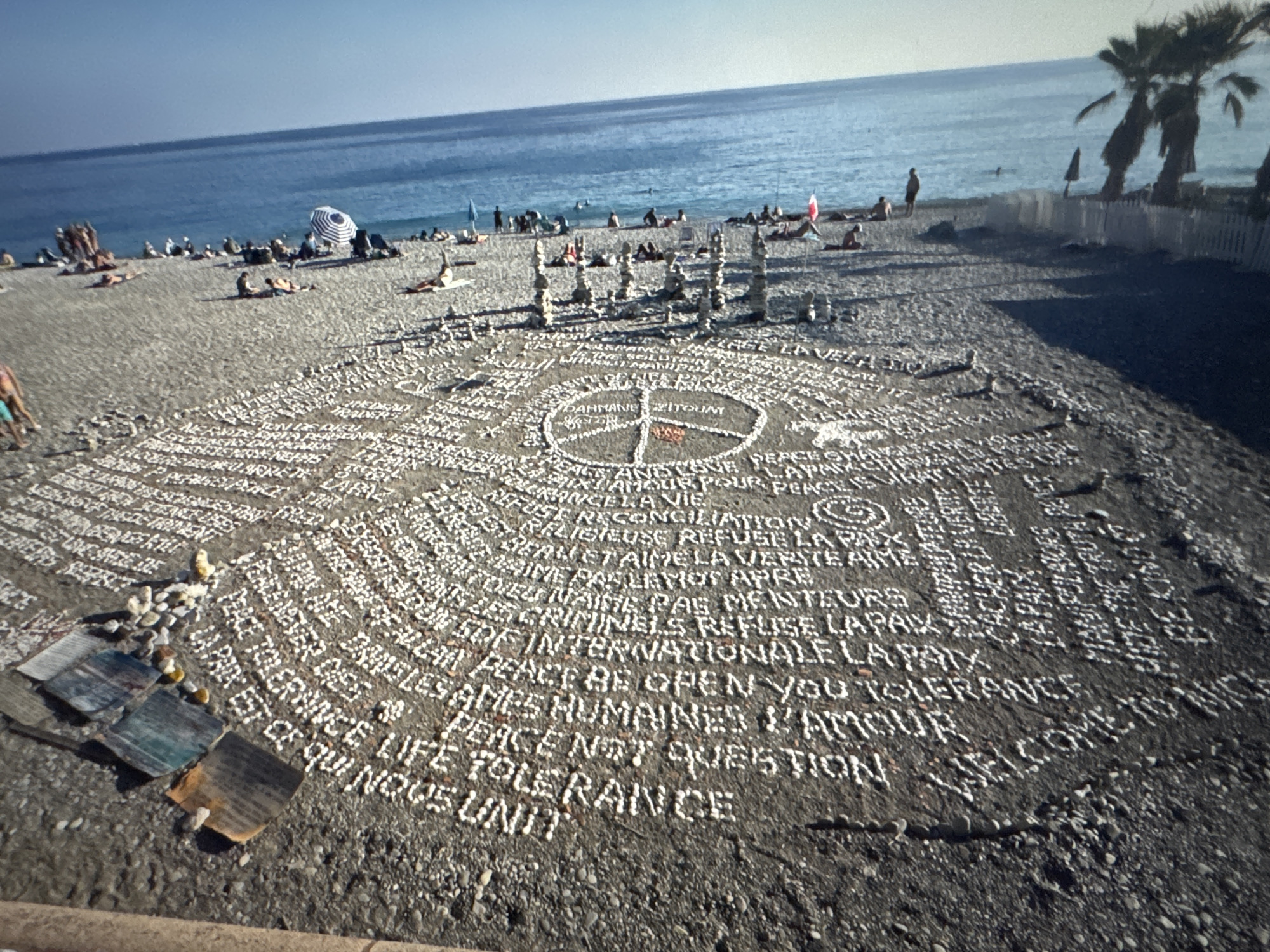 The image depicts a beach scene with a large circular arrangement of white stones on the sand, forming text. This artwork is centrally located in the frame. The text appears to be a collection of phrases related to peace, love, and tolerance, with some phrases in English and some in French. In the center of the circle is a symbol that resembles a peace sign or a compass rose.
In the background, the blue sea stretches to the horizon. The beach is populated with several people, some of whom are sunbathing or sitting on towels. Beach umbrellas and chairs are visible. There are also several tall, thin stacks of stones placed on the beach, some of which are within or near the circular text arrangement. Palm trees are visible on the right side of the image, and a white picket fence runs along the edge of the beach.
The location is identified as the city of Porrentruy, Switzerland. However, the image clearly shows a beach with a sea, which is not characteristic of Porrentruy, a landlocked city in Switzerland. The scene is more consistent with a coastal location.