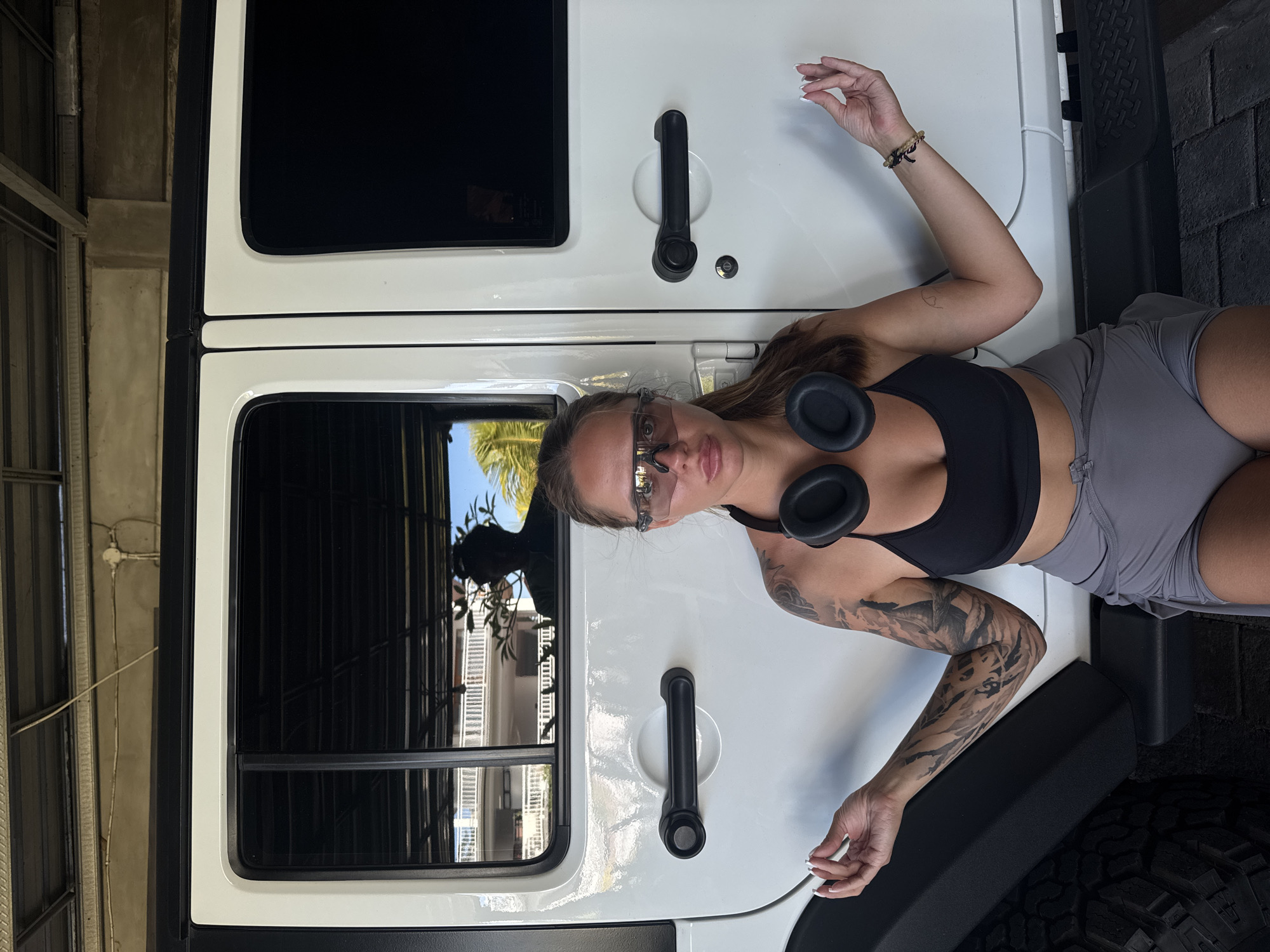 A woman lies atop the white hood of a Jeep Wrangler parked outdoors. She wears a black sports bra, gray shorts, and futuristic translucent sunglasses, and headphones are draped around her neck. Her left arm has a detailed tattoo sleeve. The Jeep has black door handles, windows, and fender flares. The location appears to be North Kuta, Indonesia, based on the architecture visible in the reflections of the window.