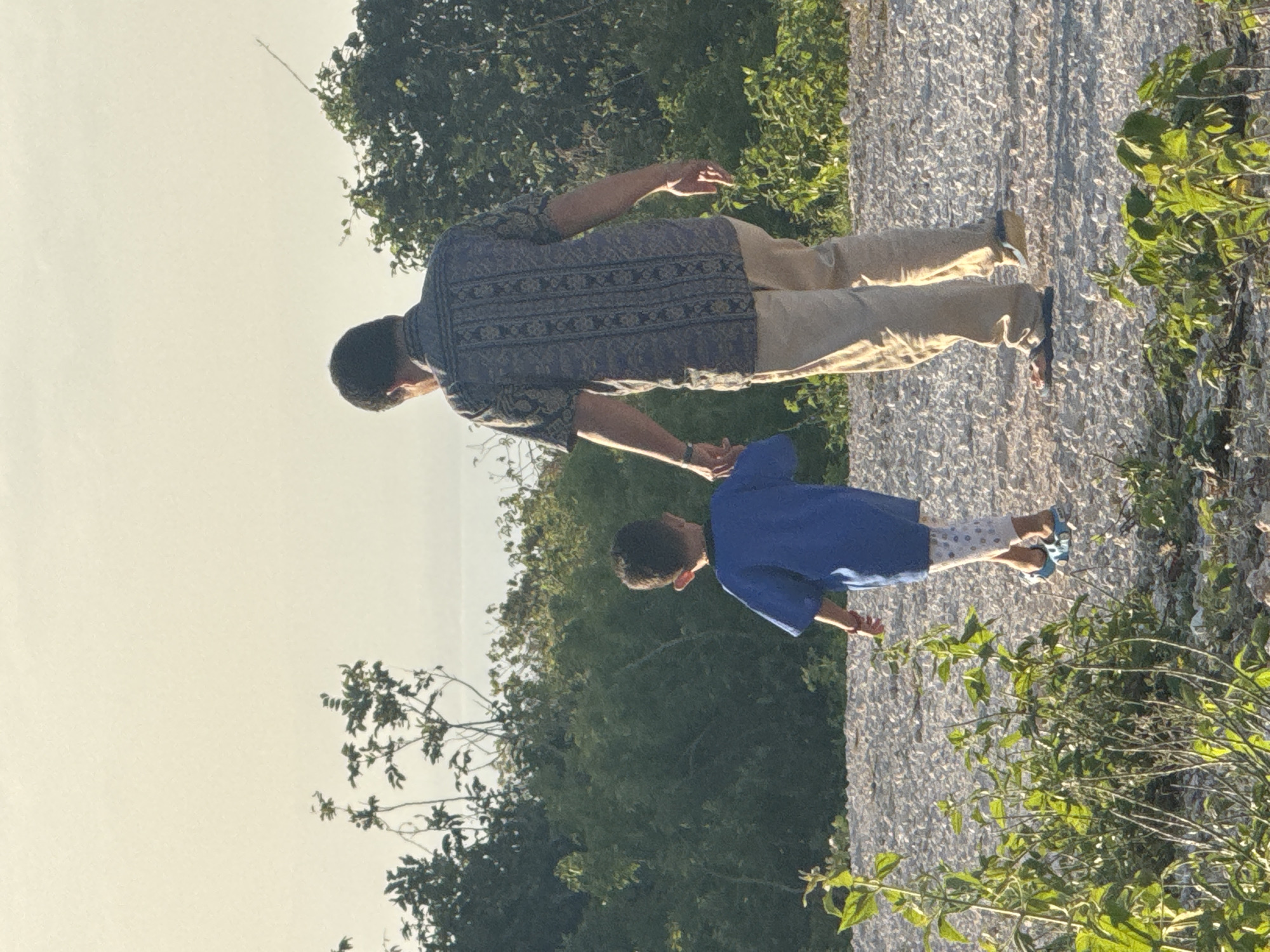 A man and a young child are walking hand-in-hand on a gravel path in Kuta Selatan, Indonesia. The man, from behind, is wearing a patterned short-sleeved shirt and tan pants. The child, also from behind, is wearing a blue t-shirt, patterned leggings, and blue sandals. They are surrounded by lush green vegetation and trees. The sky above is pale and hazy, suggesting it might be early morning or late afternoon. The ground is covered with light-colored gravel and some scattered leaves. In the distance, a body of water is visible. The overall scene depicts a peaceful walk through a natural environment.