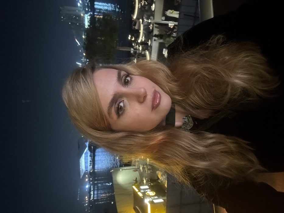 A young woman poses close to the camera at night with a city and body of water in the background. She has light blonde hair and is wearing a black top with a black choker featuring a decorative embellishment. The background showcases city lights reflected in the water, suggesting a waterfront location. The setting appears to be a restaurant or bar with outdoor seating.