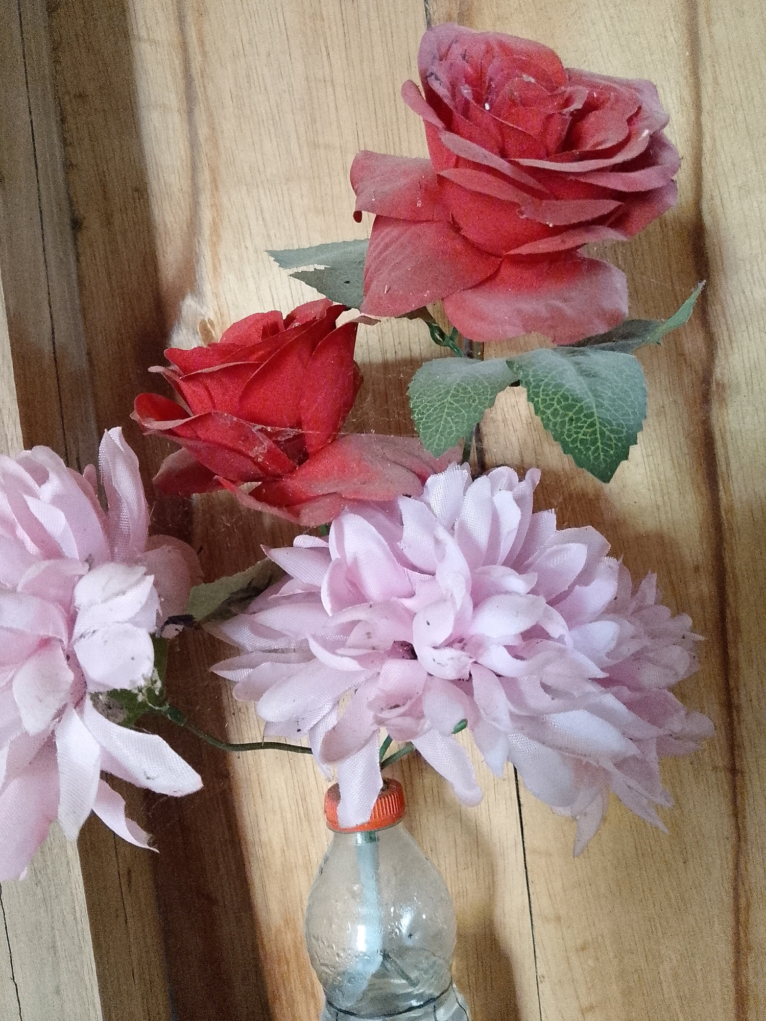 The image shows a close-up of artificial flowers in a vase against a wooden background. There are two red roses and several pink chrysanthemum-like flowers. The flowers appear to be dusty. The vase is a clear plastic bottle with an orange cap. The wooden background consists of vertical planks. There are no people or animals present in the image. The scene is indoors. There is no information in the image to determine the location within the city of Wadang, Indonesia.