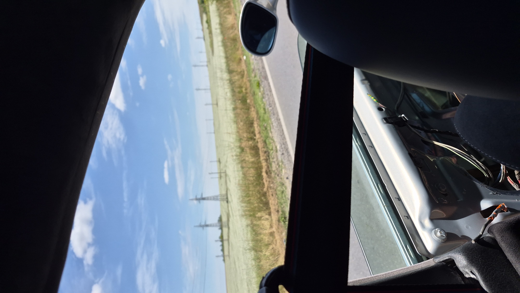 The image, captured from inside a vehicle, displays a view through an open car door. The scene outside is dominated by a bright blue sky with scattered white clouds, suggesting a sunny day. A vast, green and yellow field stretches into the distance, punctuated by several tall power line towers, indicating a rural or semi-rural landscape. A paved road runs alongside the field, with a thin strip of gravel bordering it.
The interior of the car is partially visible. The door panel has been removed, revealing exposed wiring and the car's internal structure. A dark seat is visible in the upper right portion of the frame. A side-view mirror is mounted on the exterior of the car, reflecting the passing scenery. The overall impression is of a journey on a clear day, with the interior of the car undergoing some form of maintenance or modification. No people are visible, and no text is present in the image. The lighting suggests it is daytime.