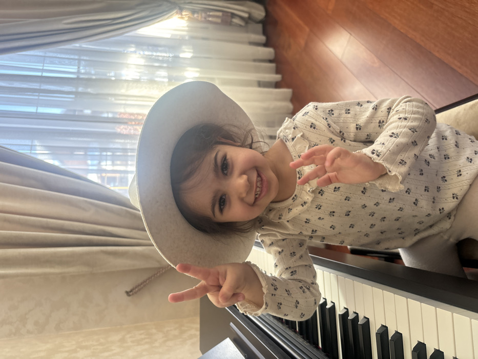 A young girl wearing a hat sits at a piano, smiling broadly and holding up her hands in a peace sign and an "OK" gesture. She wears a long-sleeved, cream-colored shirt with a small floral pattern. Sunlight streams in from a window behind her, illuminating the curtains. The piano keys are prominently visible in the foreground. The photo has been rotated slightly, giving it a tilted perspective.