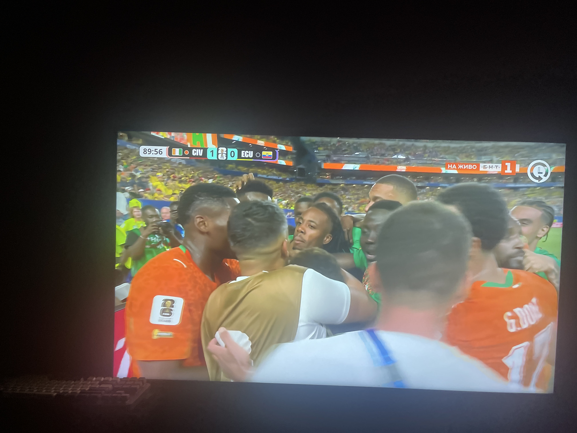 The image shows a television screen displaying a soccer match. The score is visible as "89:56 1-0 CIV ECU". Several people are gathered in front of the screen, looking at it. The screen shows a close-up of soccer players in orange and white jerseys. The background of the screen shows a stadium filled with spectators. The image is taken from a slightly elevated angle, with the television screen filling most of the frame. There is a keyboard in the lower left corner of the image. The city of Ruse, Bulgaria is not depicted in this image.