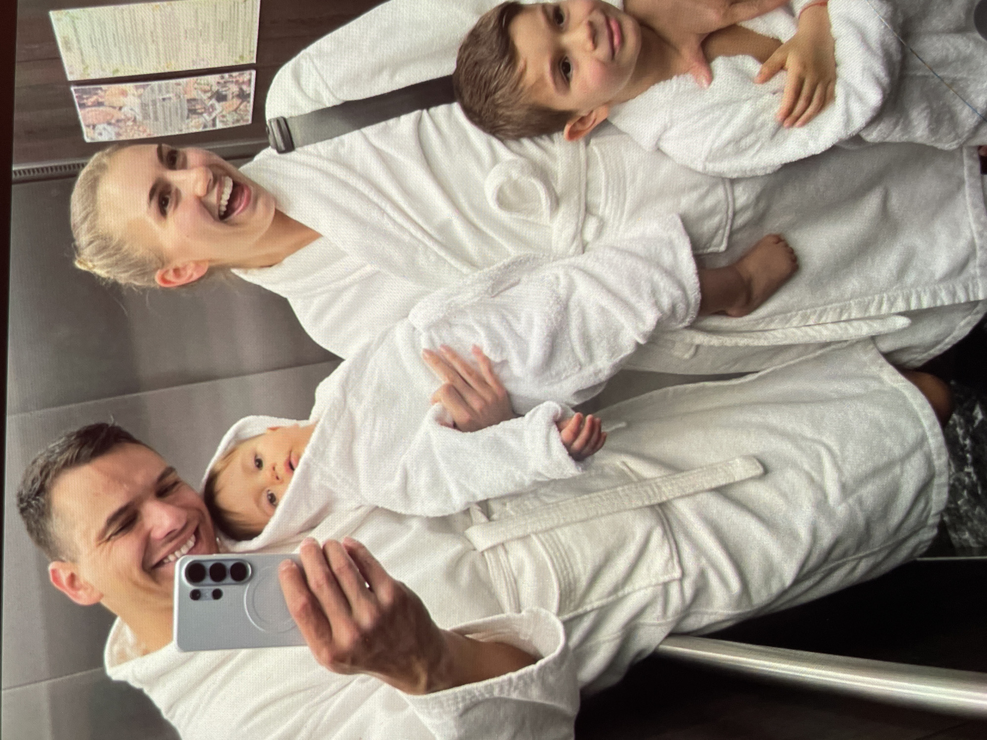 A family of four is taking a selfie in an elevator. The mother, a blonde woman with her hair pulled back, is smiling and looking upwards. The father, a man with short dark hair, is holding a silver smartphone and smiling towards the camera. They are both wearing white bathrobes. A young boy is lying on top of the mother, also wearing a white bathrobe. A baby with fair skin is nestled in the father's arms, wrapped in a white bathrobe hood. The elevator walls appear to be silver. There is a piece of paper affixed to the wall. The location is stated to be Harmanli, Bulgaria.