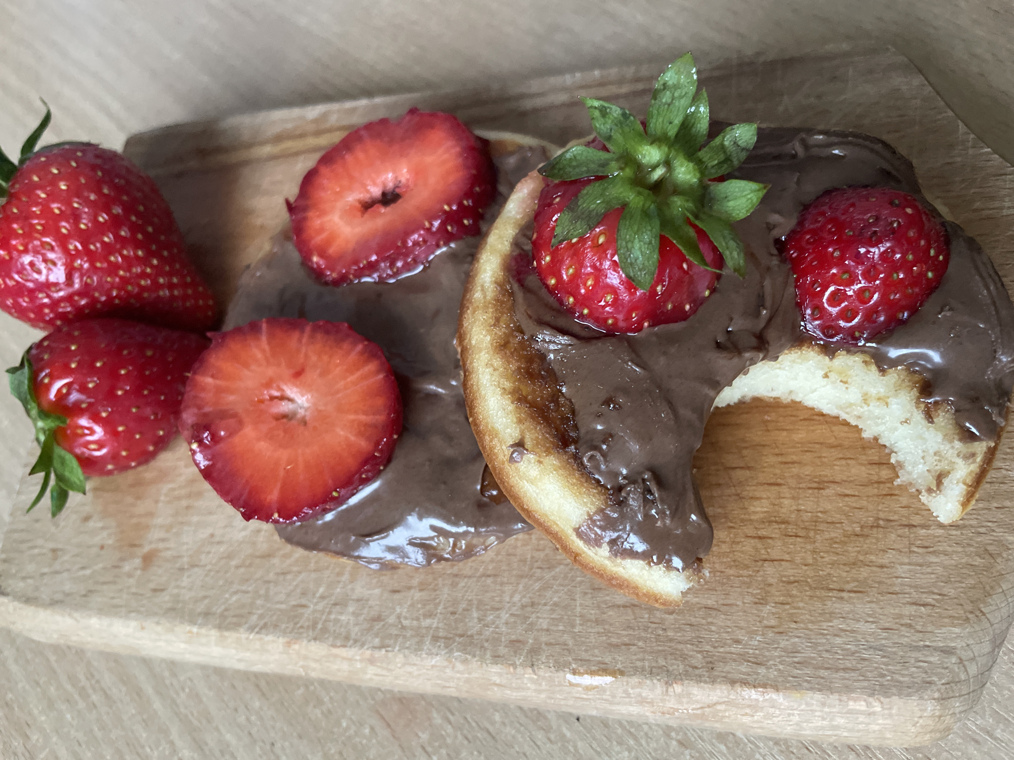 The image displays a close-up, high-angle view of three pancakes or waffles topped with chocolate spread and fresh strawberries, arranged on a light-colored wooden cutting board.
The main subjects are the food items: round, golden-brown pancakes or waffles, each generously coated with a rich, dark brown chocolate spread. Adorning the pancakes are several bright red strawberries; some are whole with their green stems intact, while others are sliced in half, revealing their juicy interiors. On the right side of the board, one of the pancakes has a noticeable bite taken out of it, exposing its light, fluffy texture beneath the chocolate.
The food is placed on a light wooden cutting board, which shows natural grain patterns and some minor surface wear. The background is softly blurred, hinting at an indoor setting, likely a kitchen or dining area. The lighting is bright and even, suggesting daytime conditions. The overall presentation indicates a prepared meal or snack, possibly breakfast or brunch, ready for consumption or in the process of being eaten. No text is visible in the image. This scene is located in Plovdiv, Bulgaria.