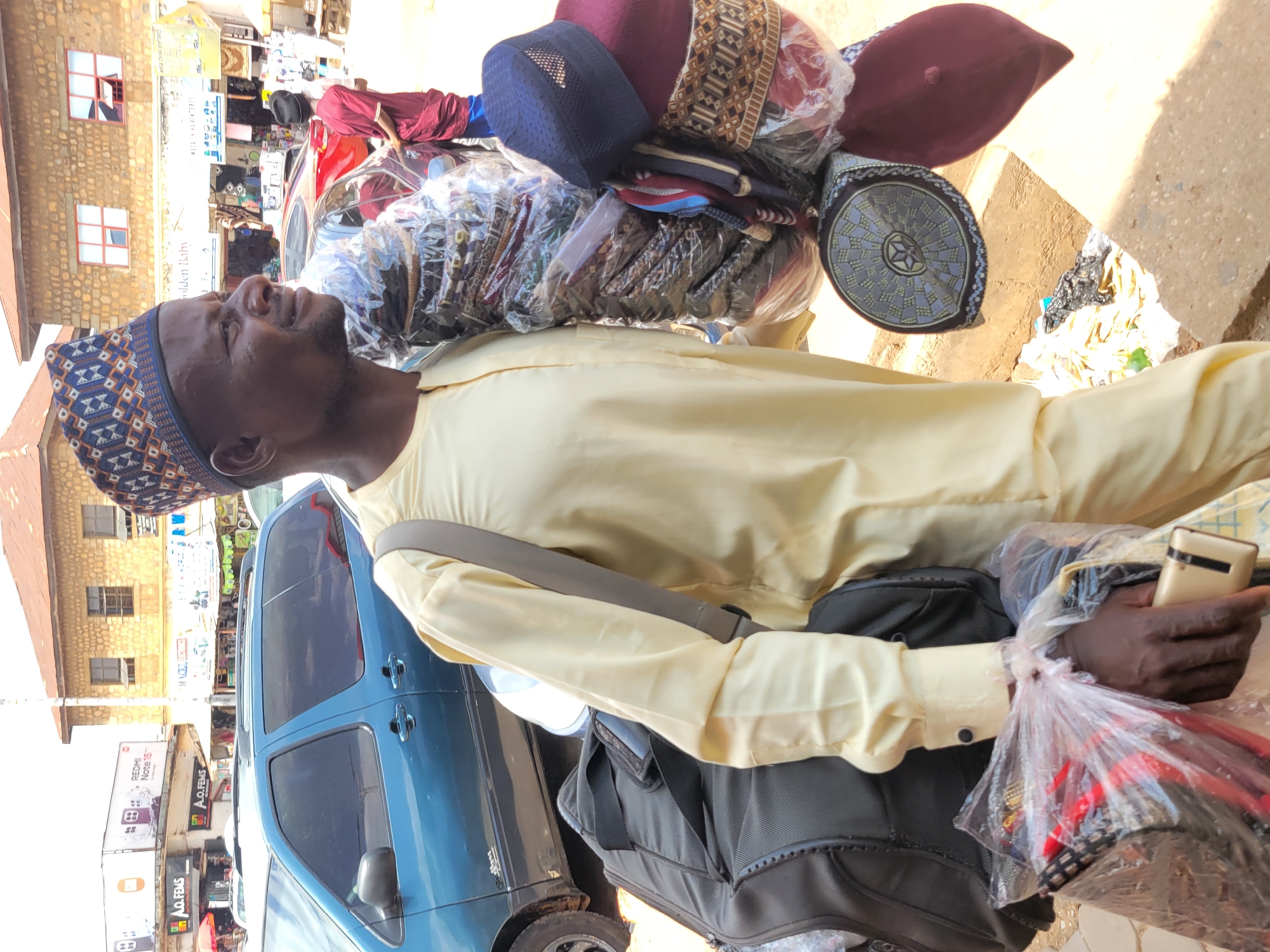 An adult male is carrying a large assortment of hats on his back and shoulder while standing outdoors on a city street in Jos, Nigeria. He wears a patterned cap, a light yellow long-sleeved shirt, and a dark cross-body bag. In his right hand, he holds a gold-colored mobile phone and a plastic bag containing more hats. The hats on his back include a variety of colors, patterns, and styles. Behind him is a blue minivan and a building with stone walls and visible windows. Street signs and advertisements are visible in the background, indicative of a commercial area. The scene appears to be a sunny day.