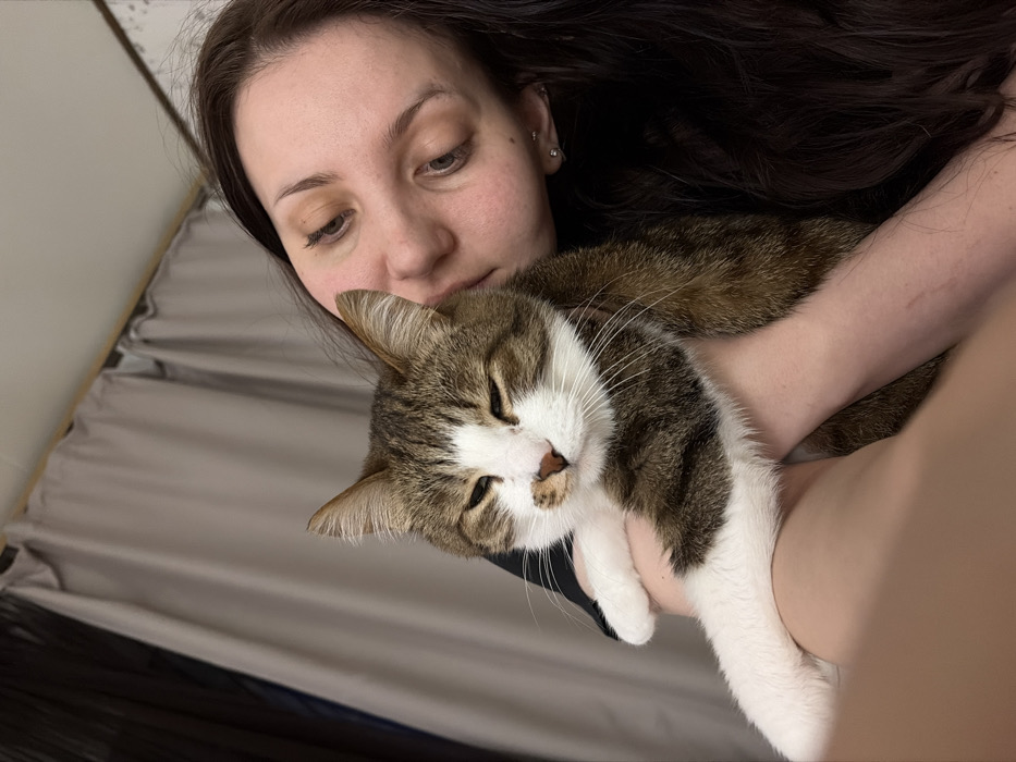 A woman with dark hair holds a tabby and white cat. She looks down at the feline, which rests comfortably in her arms with its eyes closed. The cat's white paws are prominent. The background shows a gray sheet or blanket, suggesting a bed or couch. The overall scene evokes a sense of comfort, affection, and peaceful companionship.