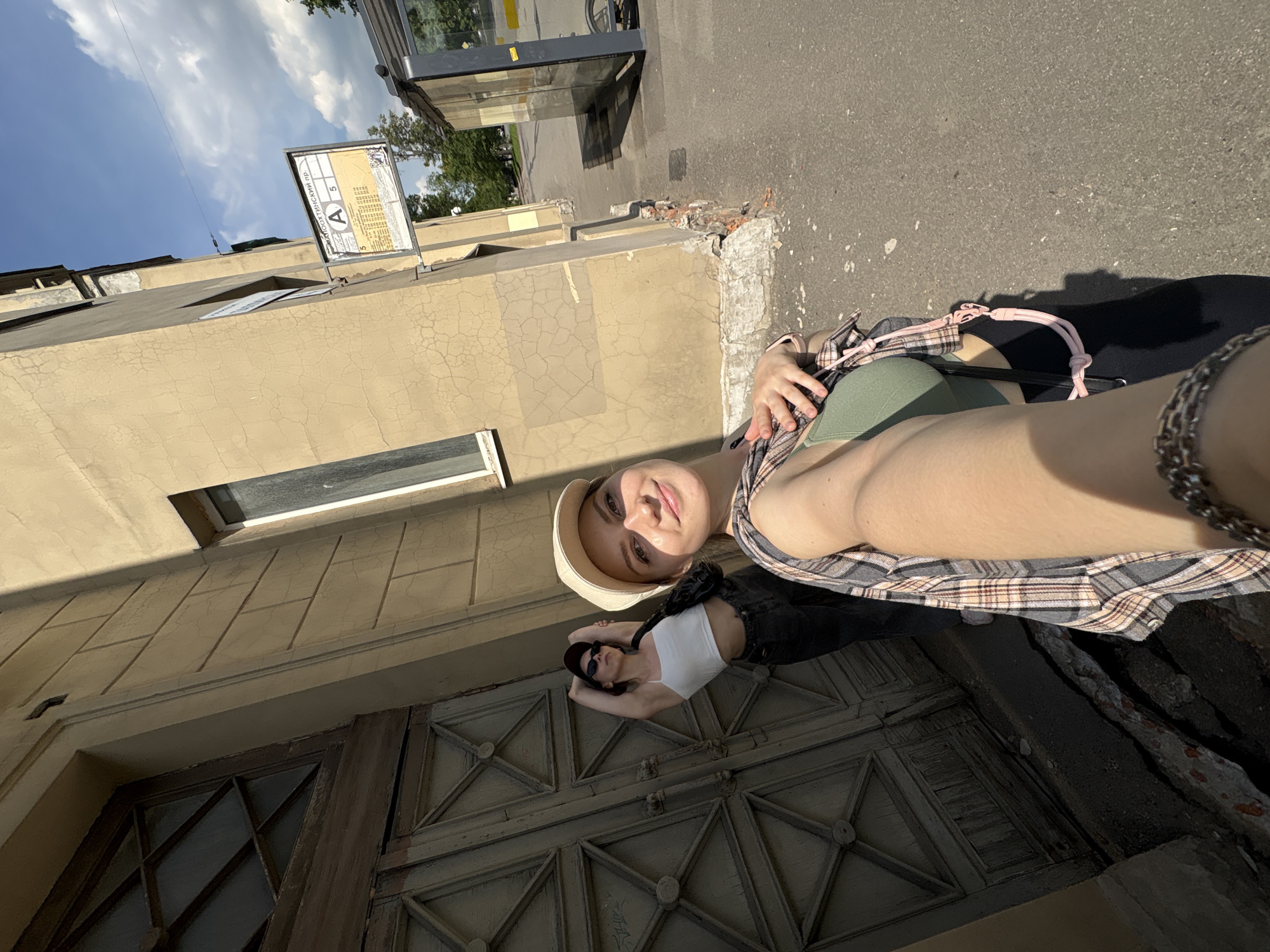 In Saint Petersburg, Russia, on a sunny day, two young women pose for a selfie in front of a weathered, tan-colored building with large, ornate wooden doors and a diamond-shaped window. The woman in the foreground, wearing a beige baseball cap, a green crop top, and a plaid shirt draped over her shoulders, smiles at the camera, her arm extended to capture the shot. Behind her, a second woman with sunglasses and a baseball cap poses with her arms behind her head. She is wearing a white tank top and dark pants. The street in front of the building is paved, and a bus stop shelter with glass panels is visible in the background, along with a sign indicating "A 5" and a bus schedule. The building's facade shows signs of age, with cracked plaster and peeling paint. The overall scene suggests a casual outing in an urban environment.