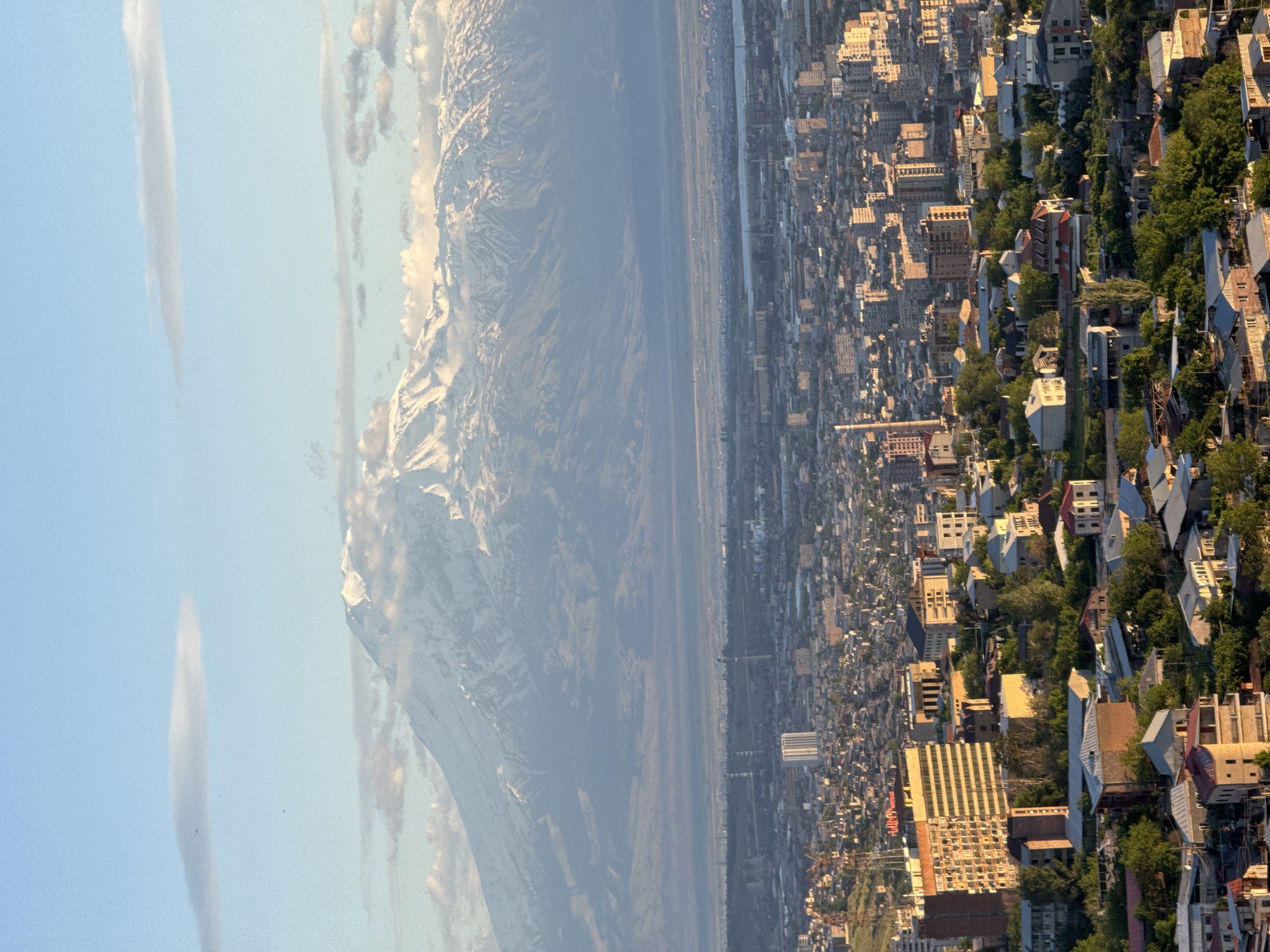 This aerial view captures a sprawling cityscape nestled against a majestic mountain range under a vast sky. The image appears to be taken from a high vantage point, looking across the city of Yerevan, Armenia, towards the iconic snow-capped peaks of Mount Ararat.
In the foreground and mid-ground, the dense urban fabric of Yerevan extends with numerous buildings of varying heights and architectural styles. Residential areas with houses and green trees intersperse with larger commercial or apartment blocks. The city appears to be arranged along a relatively flat terrain, with some gentle undulations. The lighting suggests late afternoon or early evening, as warm, golden sunlight bathes the west-facing sides of buildings, casting long shadows across the city.
A broad valley or plain separates the city from the distant mountains. In the background, the impressive, snow-covered summits of Mount Ararat dominate the horizon. Clouds cling to and hover above parts of the mountain range, adding to its grandeur. The sky above is a clear, pale blue with a few wispy, elongated clouds.
Visible text on a prominent building within the city, characterized by a red sign, clearly spells out "ARARAT" in white lettering, likely referring to the famous Armenian brandy or a related establishment, further confirming the location context. The overall scene presents a harmonious blend of urban development and natural splendor, characteristic of the Armenian capital and its unique geographical setting.