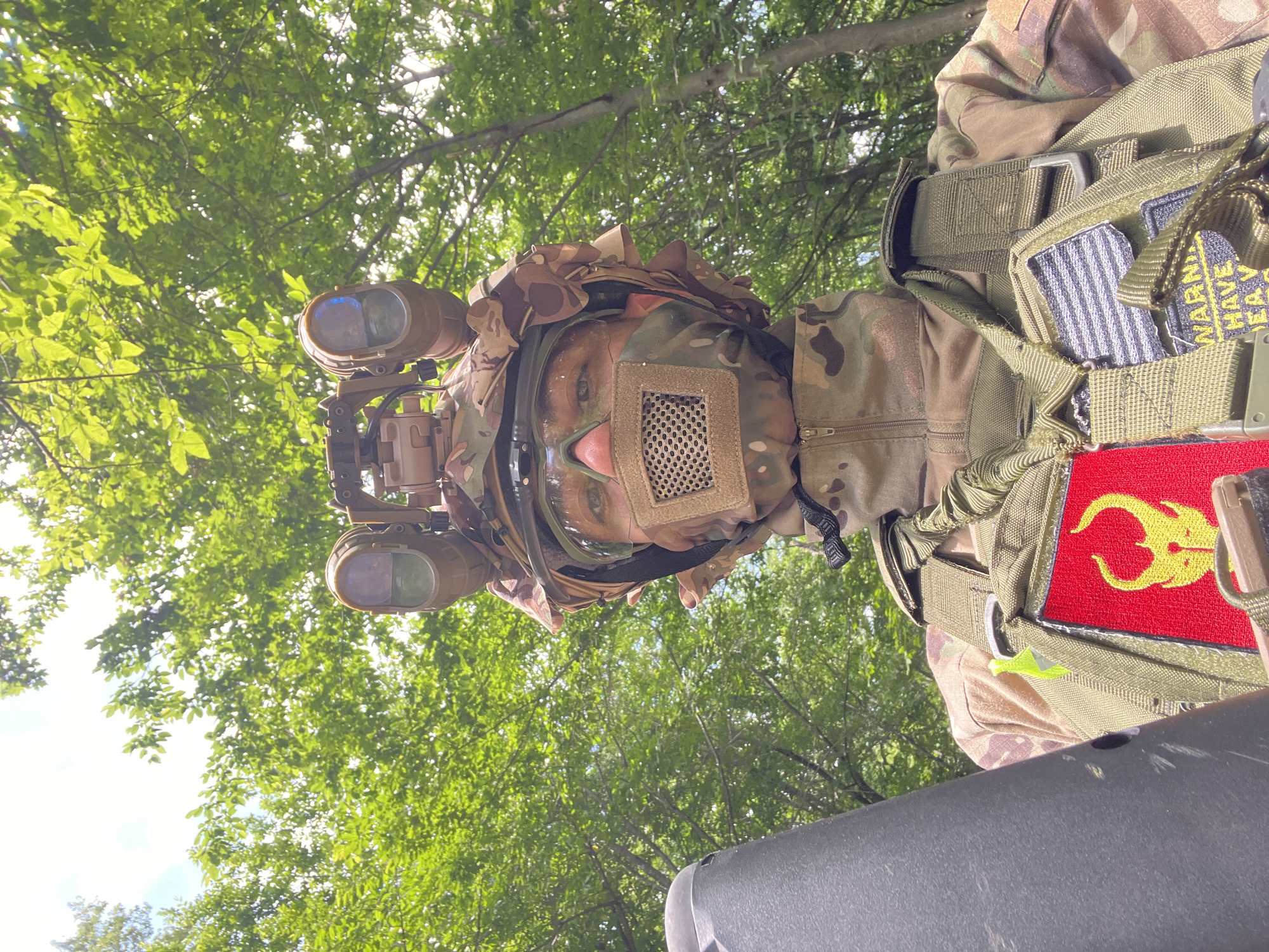 A person wearing camouflage clothing and night vision goggles is standing outside in a forested area. The person is wearing a mesh face covering and has a vest with patches on it. The leaves and sky are visible behind the person. The city is not mentioned.