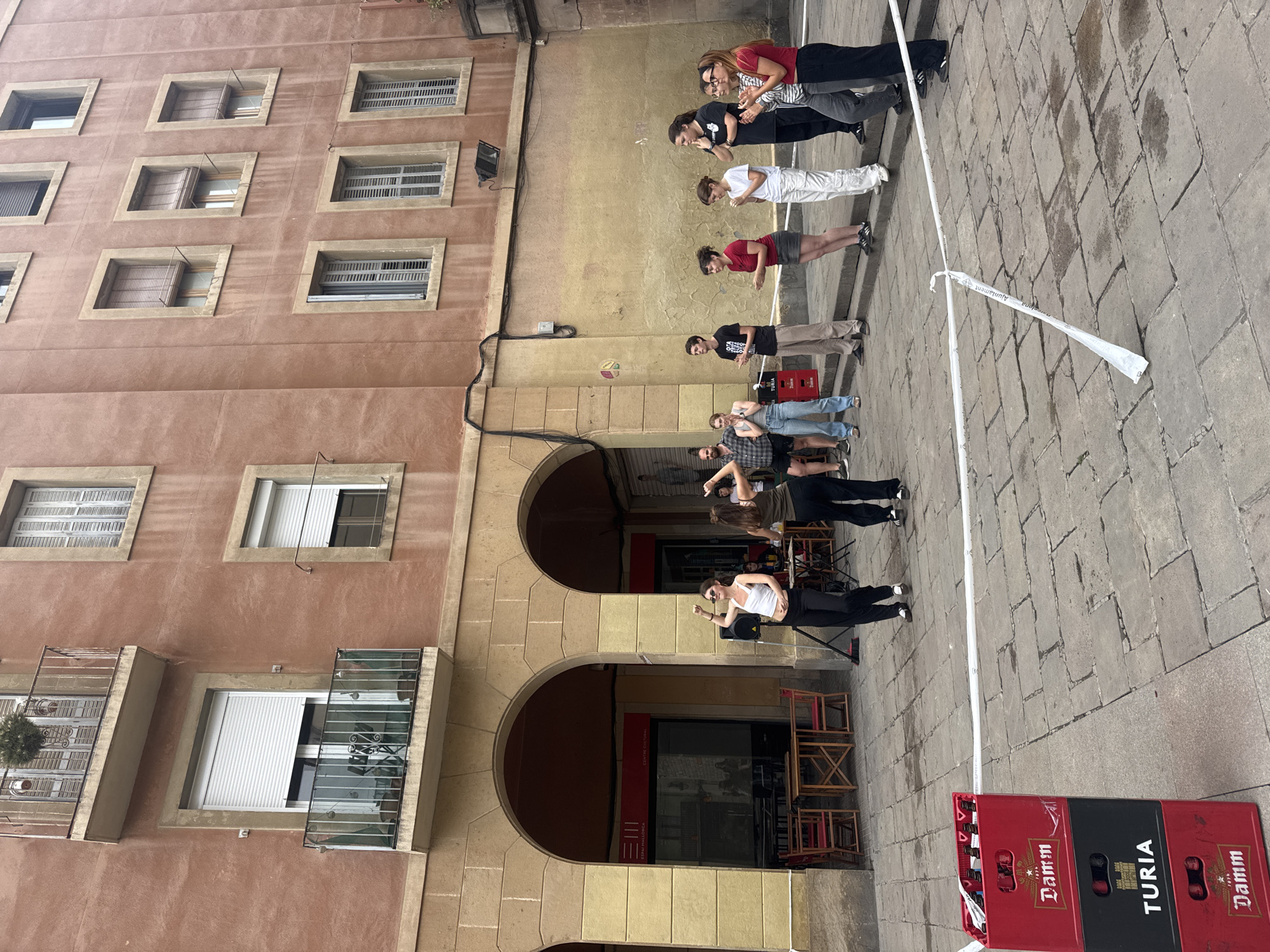A medium-shot photo shows a group of people standing in a line on a cobbled street in Barcelona, Spain. The shot is taken outdoors during the day, with natural light illuminating the scene.
The group, consisting of approximately ten individuals of various heights and builds, appears to be engaged in some sort of activity or performance. They are spaced apart along a white line painted on the pavement, with their faces turned towards the ground.
Behind the group, there's an arched colonnade that leads into a building, likely a cultural center or a shop, with the sign "Espai Mallorca". To the right of the group, there are stacks of red and black beer crates of Damm and Turia beer.
The architectural backdrop consists of a large, multi-story building with a salmon-colored facade. It features rows of rectangular windows, some with shutters or curtains. There are also a couple of small balconies with plants on them.
The scene is set within a historic urban environment, evidenced by the traditional architecture and the cobbled street. The presence of the beer crates suggests a nearby bar or restaurant. The combination of the location and the performance implies a blend of cultural activity and daily life in Barcelona.