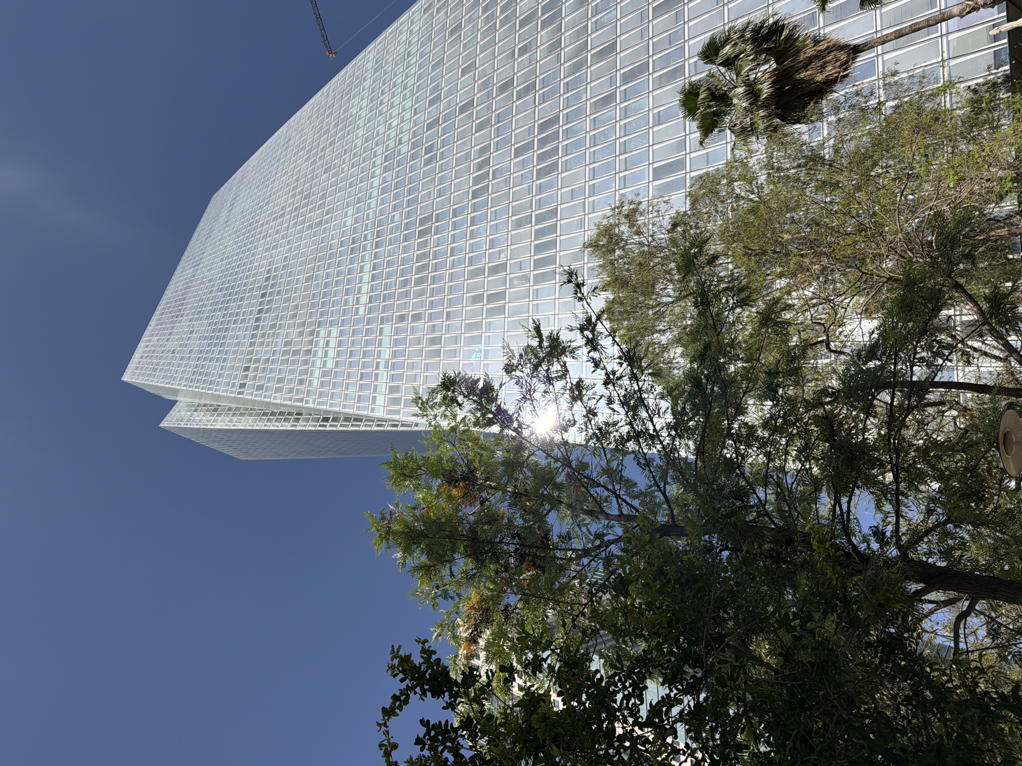 The image shows a tall, modern skyscraper in Tel Aviv, Israel. The building has a grid-like facade of glass panels that reflect the sky and surrounding structures. A construction crane is visible in the upper left corner of the image. The building is partially obscured by trees. The sky is clear and blue.