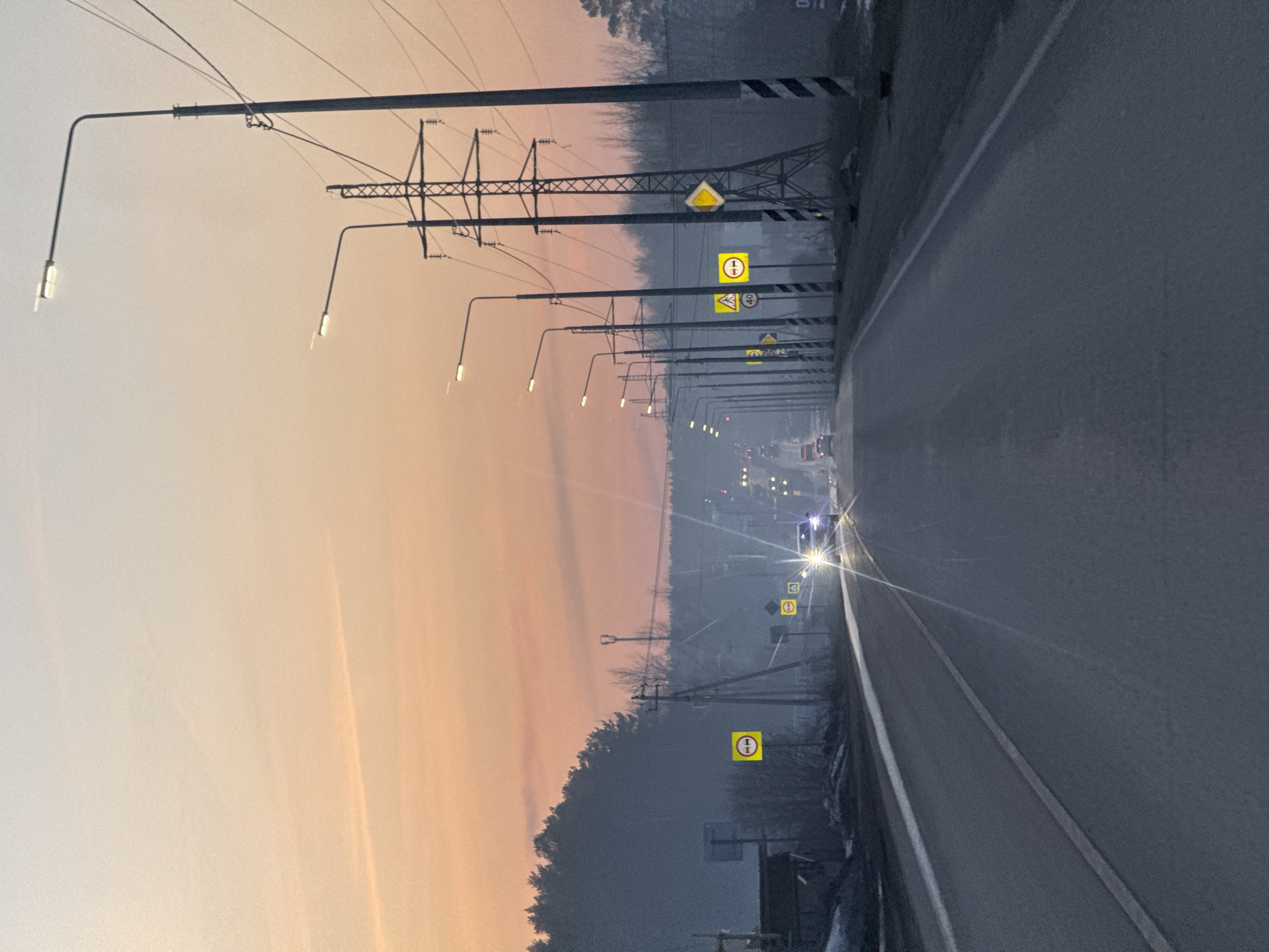 The image shows a view of a road in Chaikovsky, Russia, lined with streetlights and power lines, suggesting an industrial or suburban area. Traffic is sparse, with at least two vehicles visible in the distance. The sky displays colors associated with dawn or dusk. Various yellow road signs are visible along the side of the road.