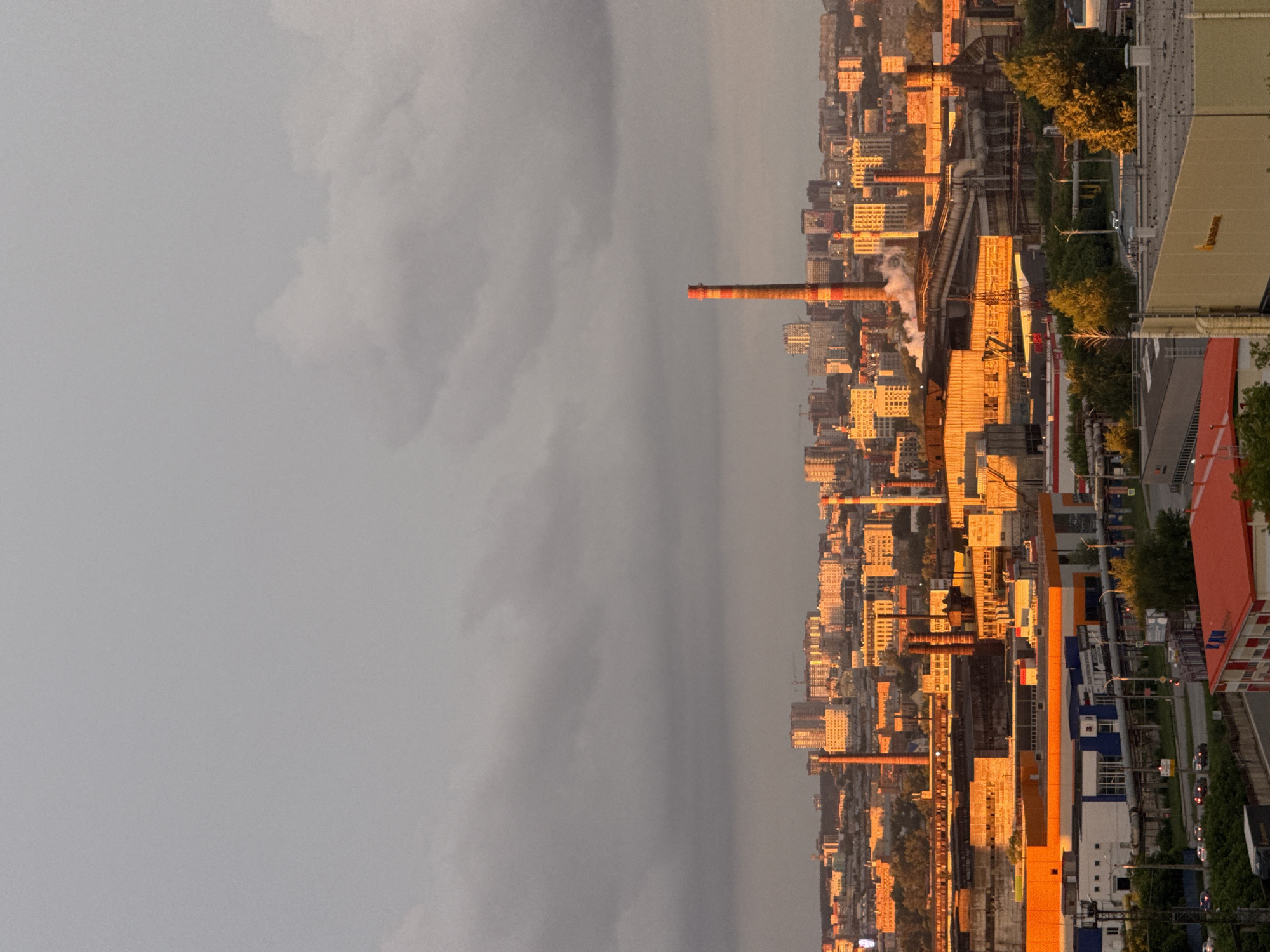 This image depicts a cityscape at sunset, with the sun's golden light casting long shadows and illuminating the buildings and industrial structures. The scene is dominated by a dense collection of multi-story buildings, likely residential and commercial, interspersed with industrial elements like smokestacks and pipes. One prominent smokestack in the foreground is orange and red striped and emits a plume of smoke, suggesting ongoing industrial activity. The sky is overcast, with darker clouds on the left suggesting approaching weather, contrasting with the warm, diffused light from the setting sun on the right.
The image appears to be taken from an elevated viewpoint, looking down on the city. The setting suggests an urban industrial area. The time of day is clearly sunset, indicated by the warm, low-angle light. The presence of clouds suggests changeable weather, possibly a storm brewing. No people are clearly visible, but the presence of cars on a road indicates it's not a deserted area. The overall mood is a blend of urban development, industrial presence, and the dramatic beauty of a sunset.
While no prominent text is legible due to image resolution and angle, the sheer density of buildings and industrial infrastructure points to a developed urban environment. The location context of Izhevsk, Russia, further grounds this industrial cityscape within a specific geographical setting.