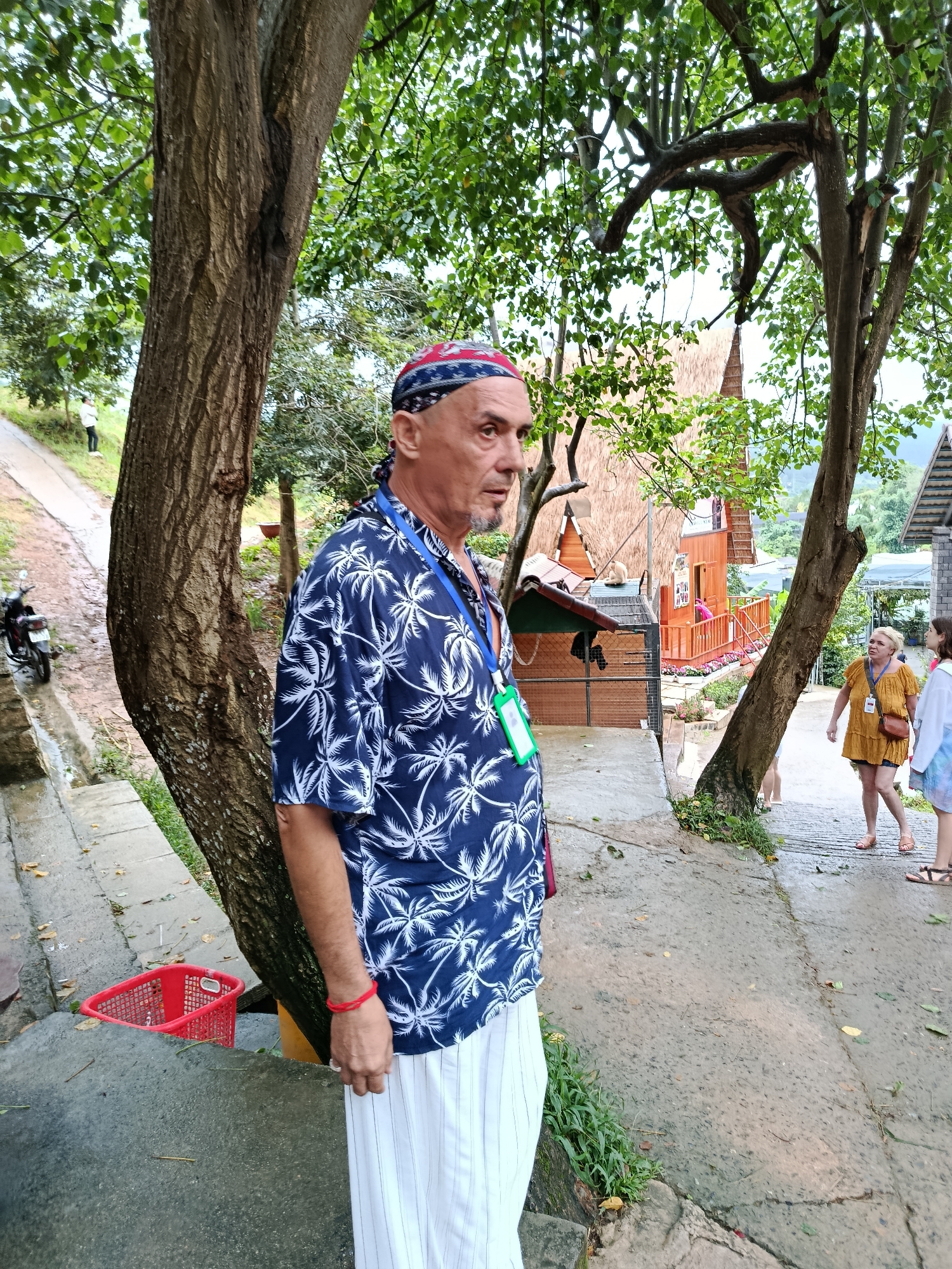 A man with a bandana and a blue short-sleeved shirt with a palm tree print stands on a wet, paved pathway. He is wearing white striped pants and a red bracelet on his left wrist. A lanyard with green and white tags is around his neck. To his left, a red basket is visible on a grey concrete ledge. In the background, there are trees, buildings with thatched roofs, and other people walking on the path. A motorcycle is parked to the left of the man. The scene appears to be in an outdoor tourist area in Định An, Vietnam.